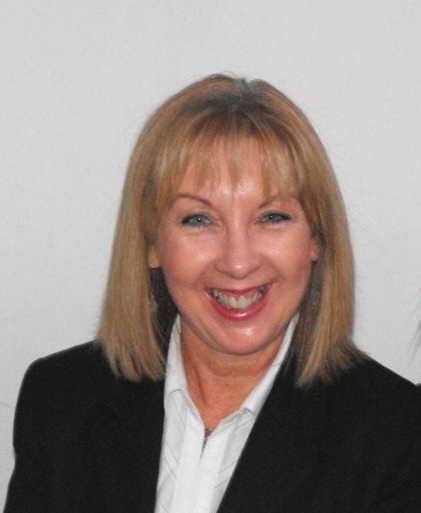 A smiling middle-aged woman with blonde hair, hoop earrings, and a blue patterned top standing against a dark gray wall.