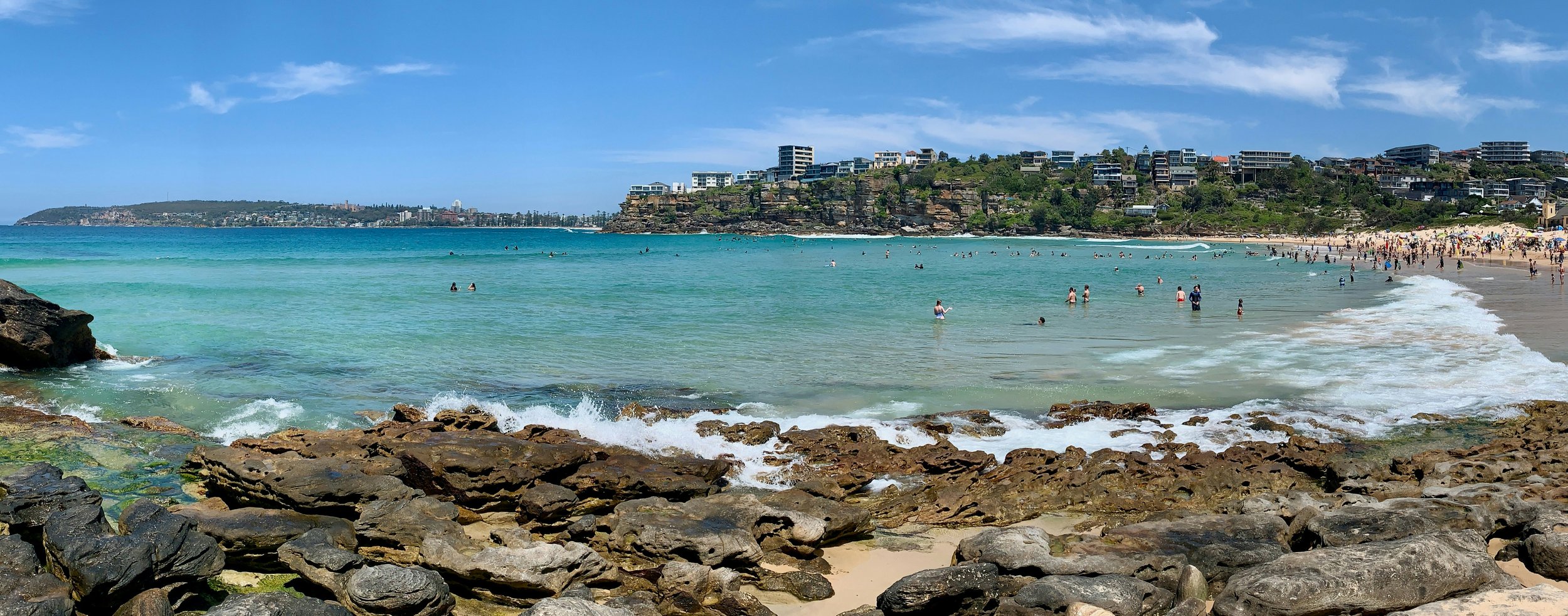 A busy beach with rocks in the foreground, clear ocean water, and a hillside with modern buildings in the background on a sunny day