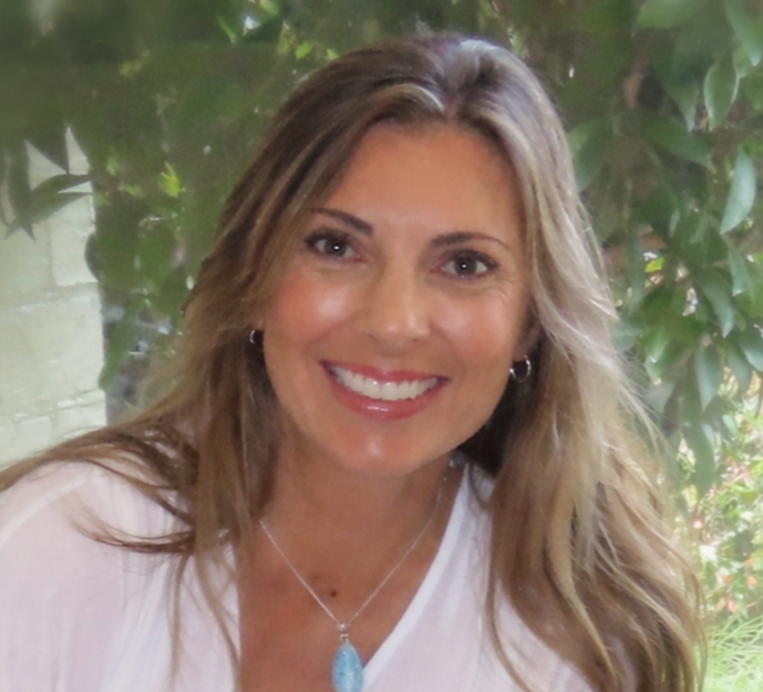 A woman with shoulder-length blonde hair smiling, wearing earrings and a black top, standing in front of a gray textured wall.