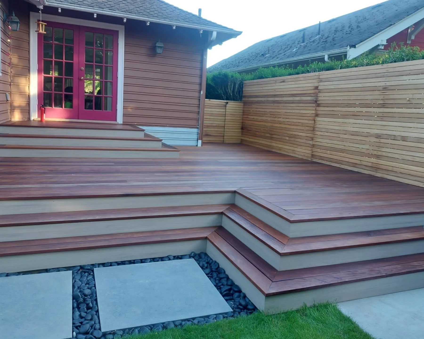 Newly constructed wooden deck outside a house with pink double doors and stairs leading up to the entrance. The area is enclosed by a wooden fence.