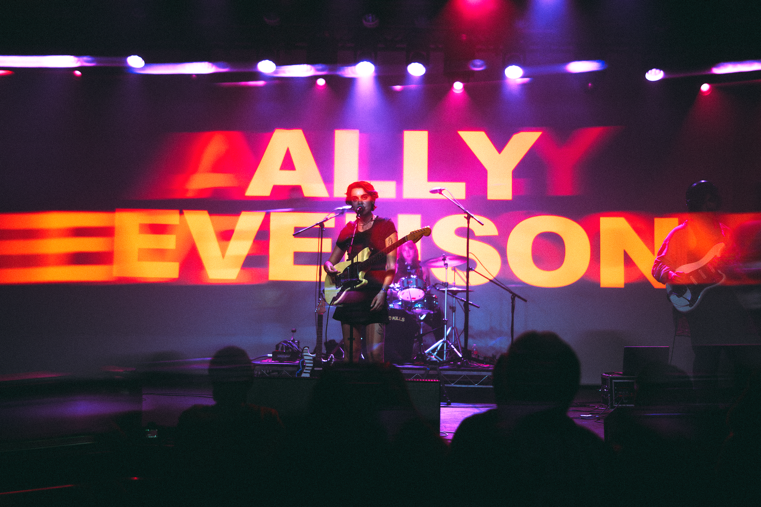 A woman performing on stage with musicians, illuminated by colored stage lights, with a large screen behind her displaying the words 'ALLY EVELSON' in red and orange text.