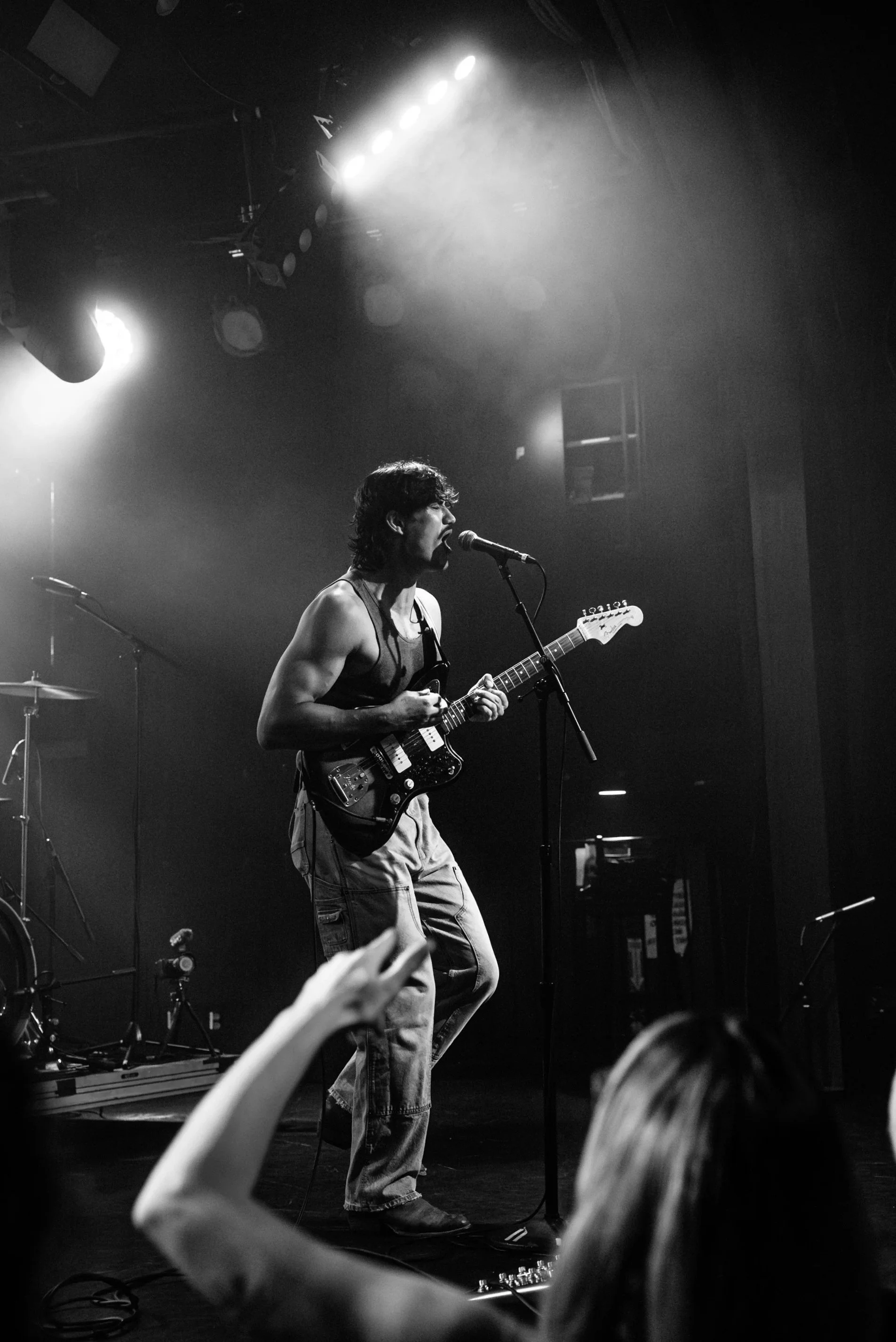 A musician playing an electric guitar and singing into a microphone on a stage. Stage lights are illuminating the scene, and a drum set and other musical equipment are visible in the background.