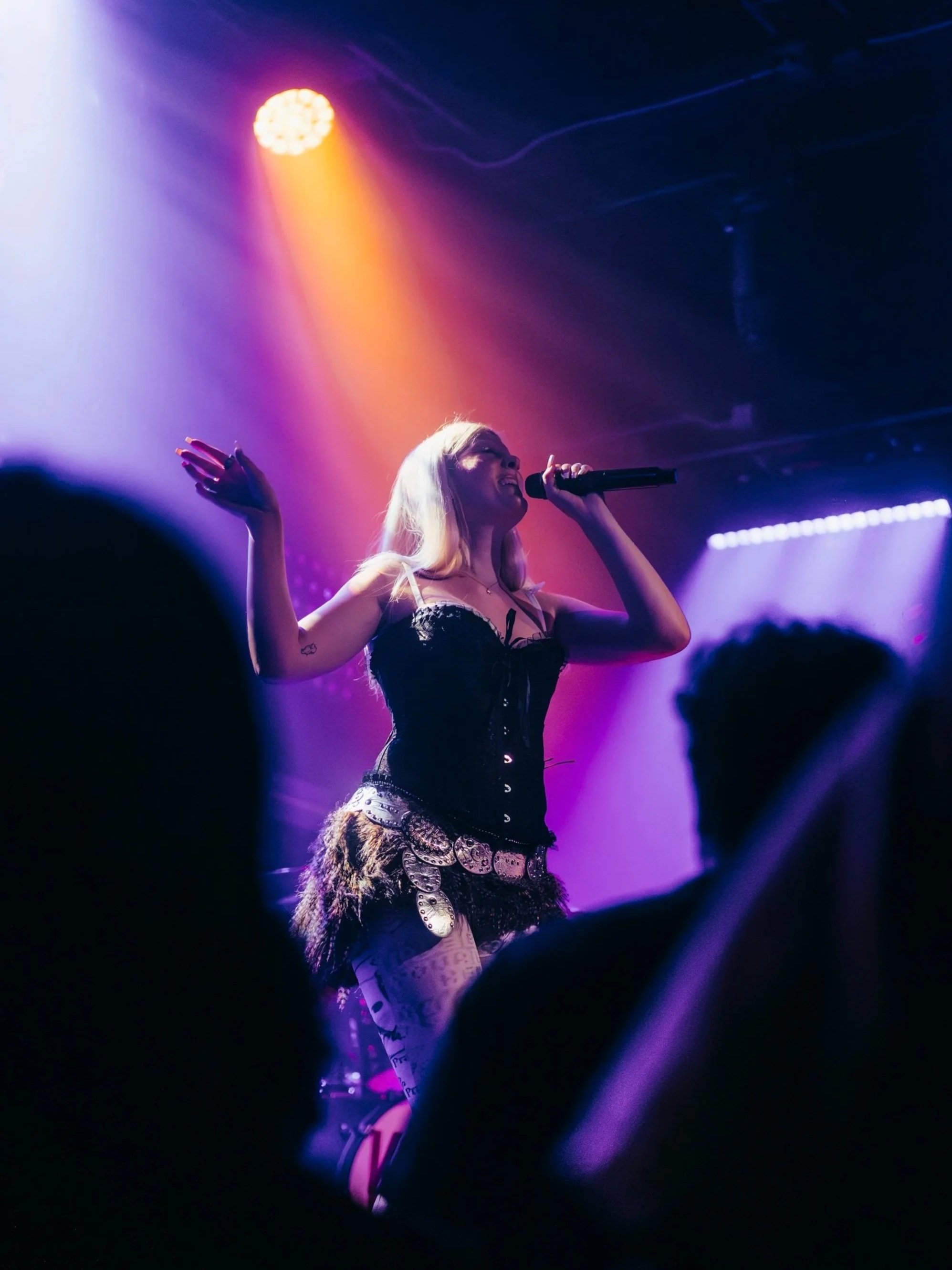 A female singer performing on stage under colorful purple, pink, and yellow lights, with a dark background and audience silhouettes visible in the foreground.