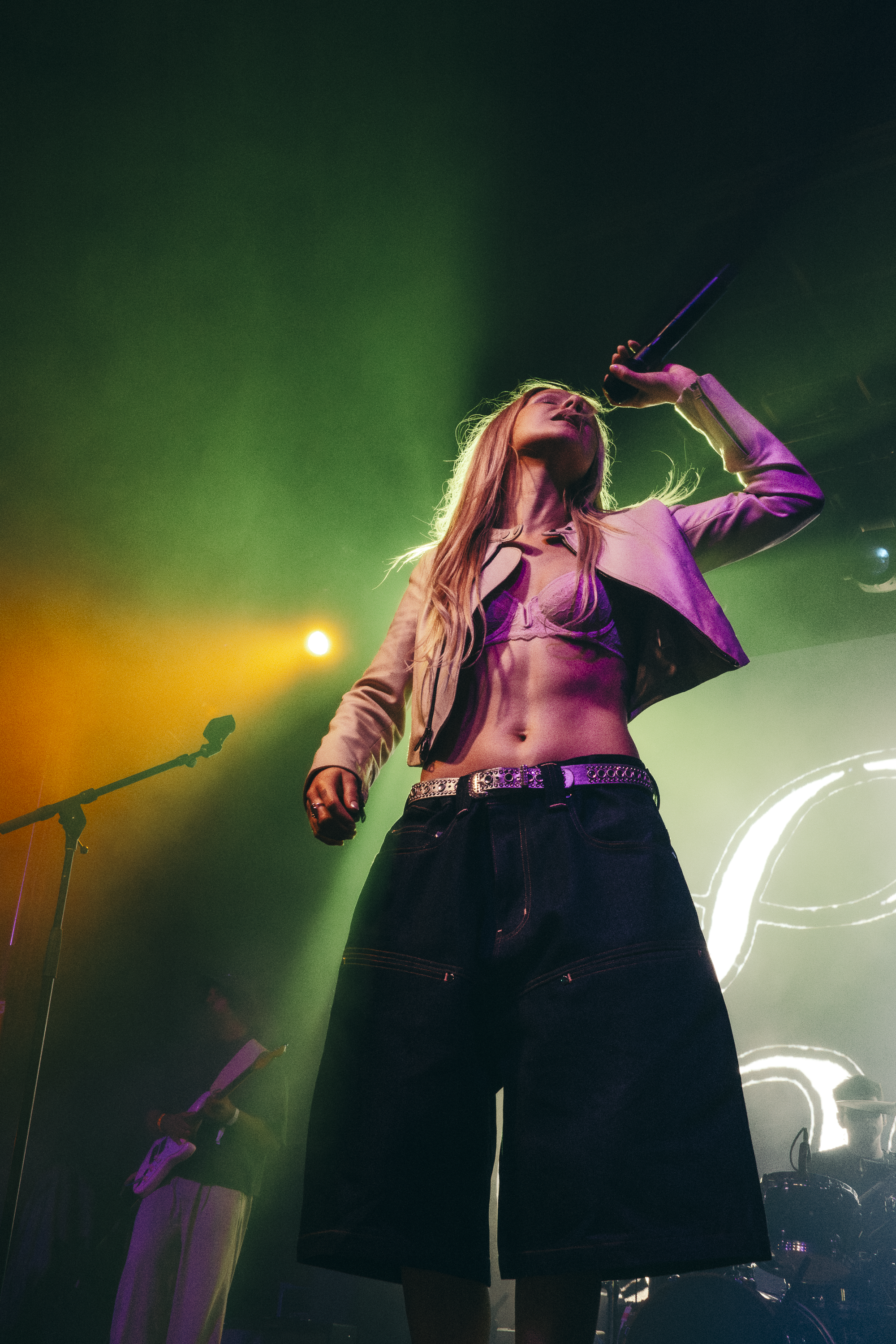 Female singer performing on stage with a microphone in her hand, wearing a cropped top, a pink jacket, and baggy jeans, with colorful stage lighting around her.