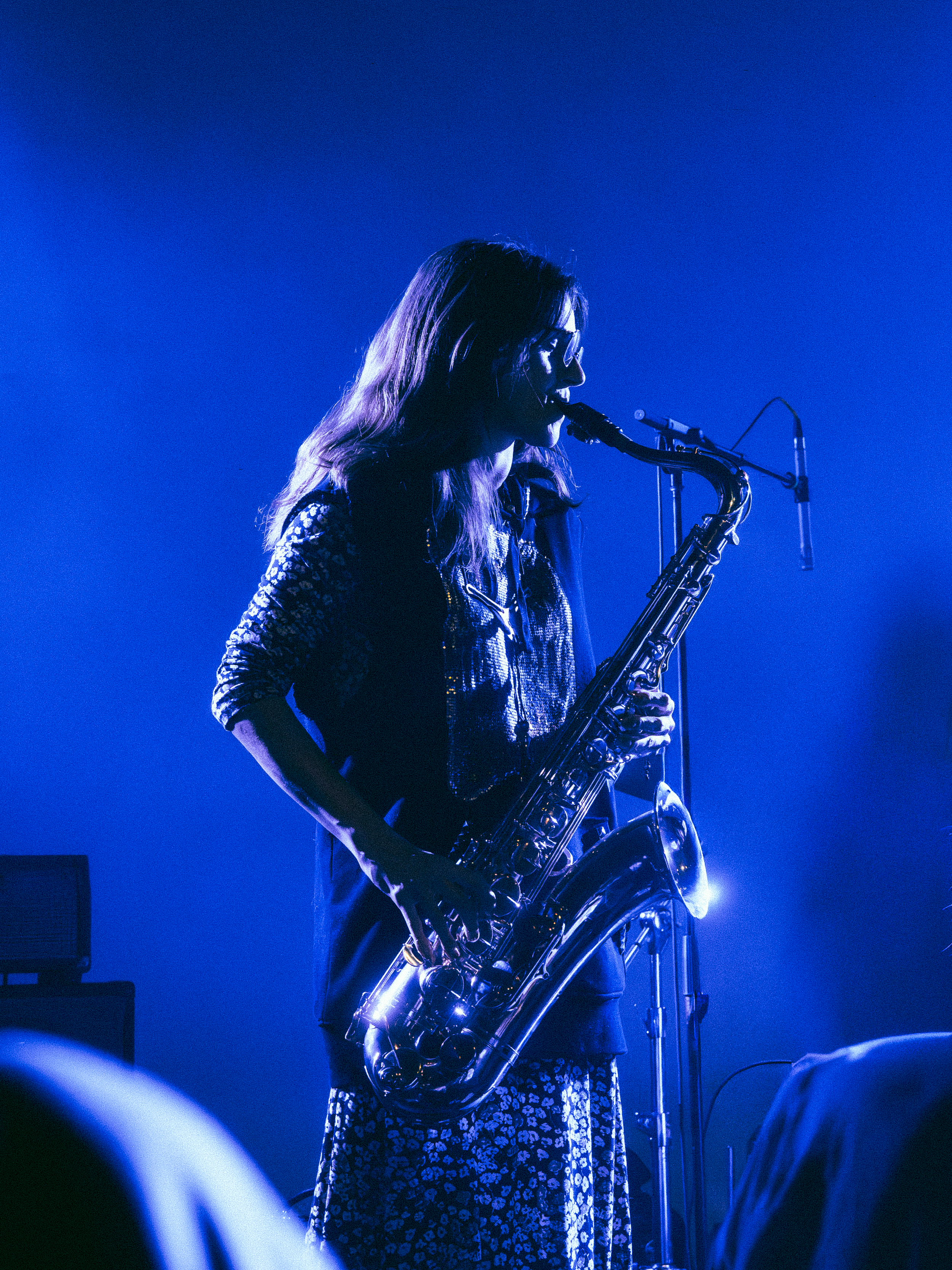 A woman wearing glasses playing a saxophone on stage, illuminated by blue stage lighting.