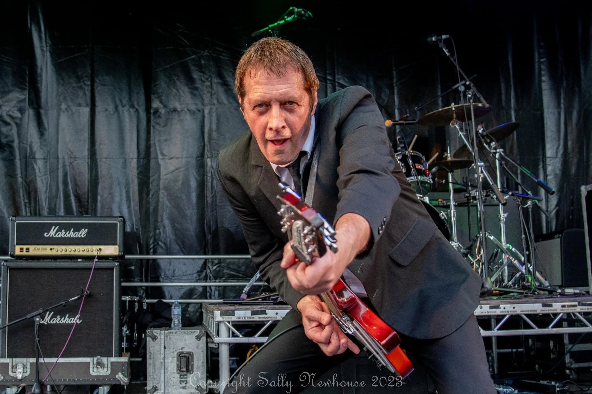Andy Coultas in a black suit and white shirt playing a Rickenbacker 330 on stage with a drum set and Marshall amplifiers in the background.