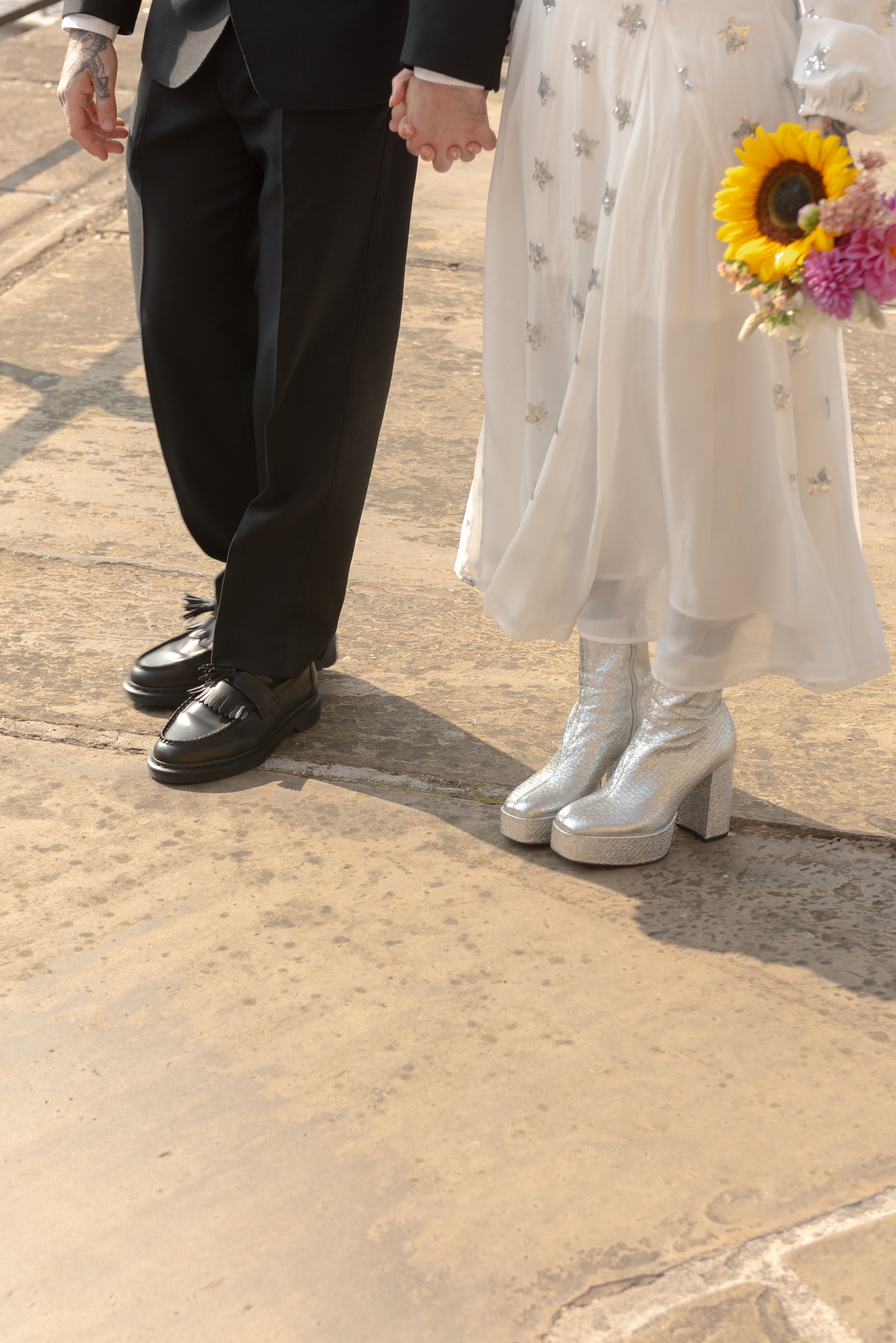 Close-up of a bride and groom holding hands during their wedding, with the bride wearing silver platform boots and the groom wearing black dress shoes, on a stone pavement.
