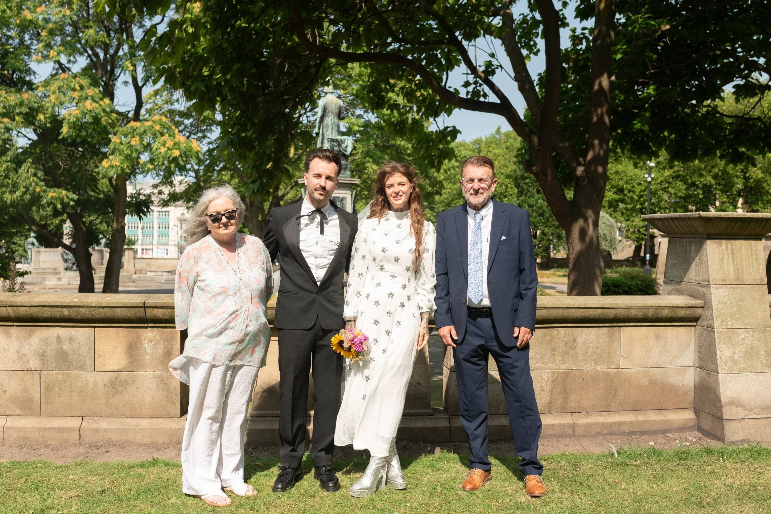 A group of five people standing outdoors in a park on a sunny day, including a bride in a white dress holding a bouquet, and four adults dressed in formal and casual attire, with trees and a statue in the background.