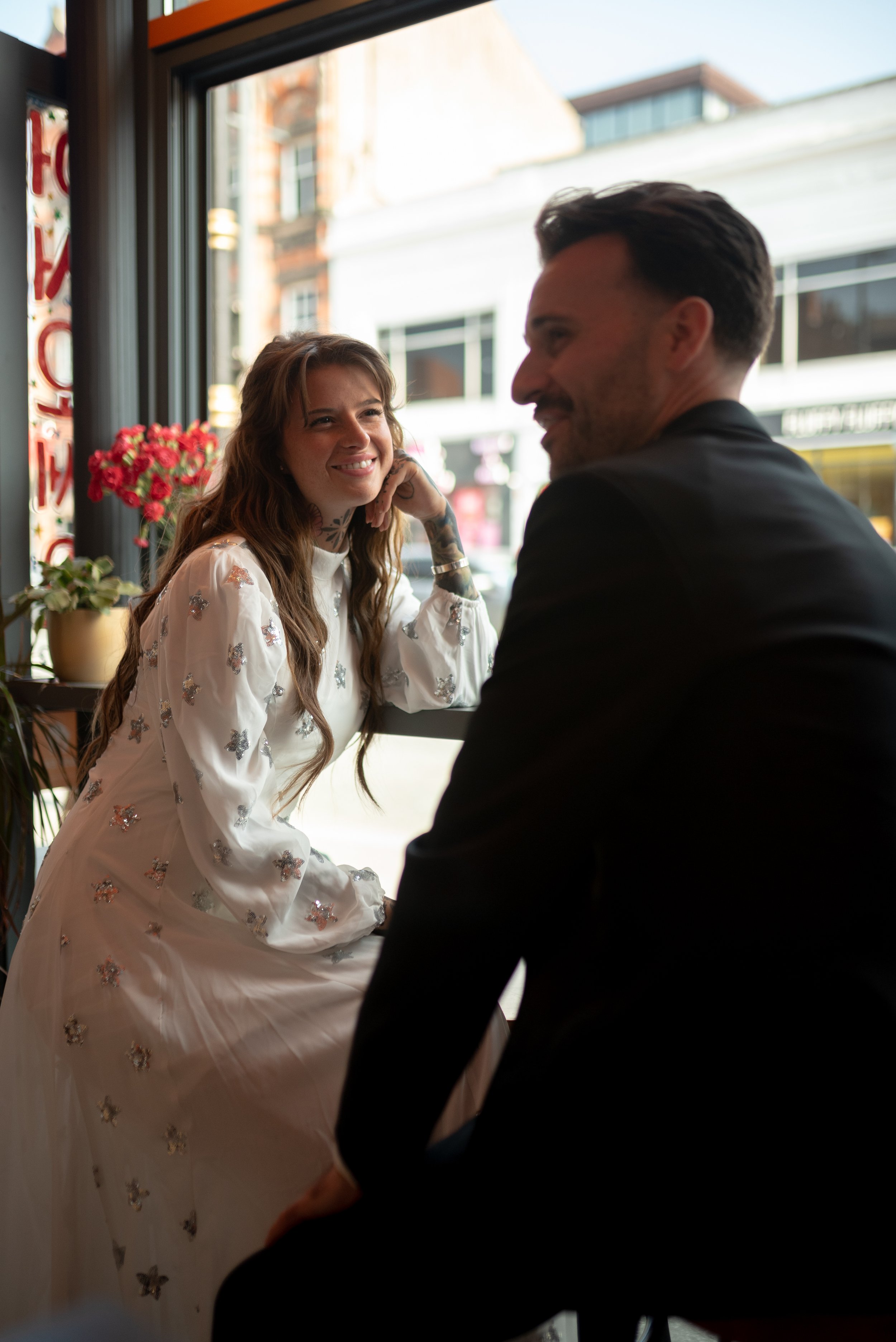 A woman with long hair and tattoos smiling at a man sitting across from her in a café near a window with city street view.