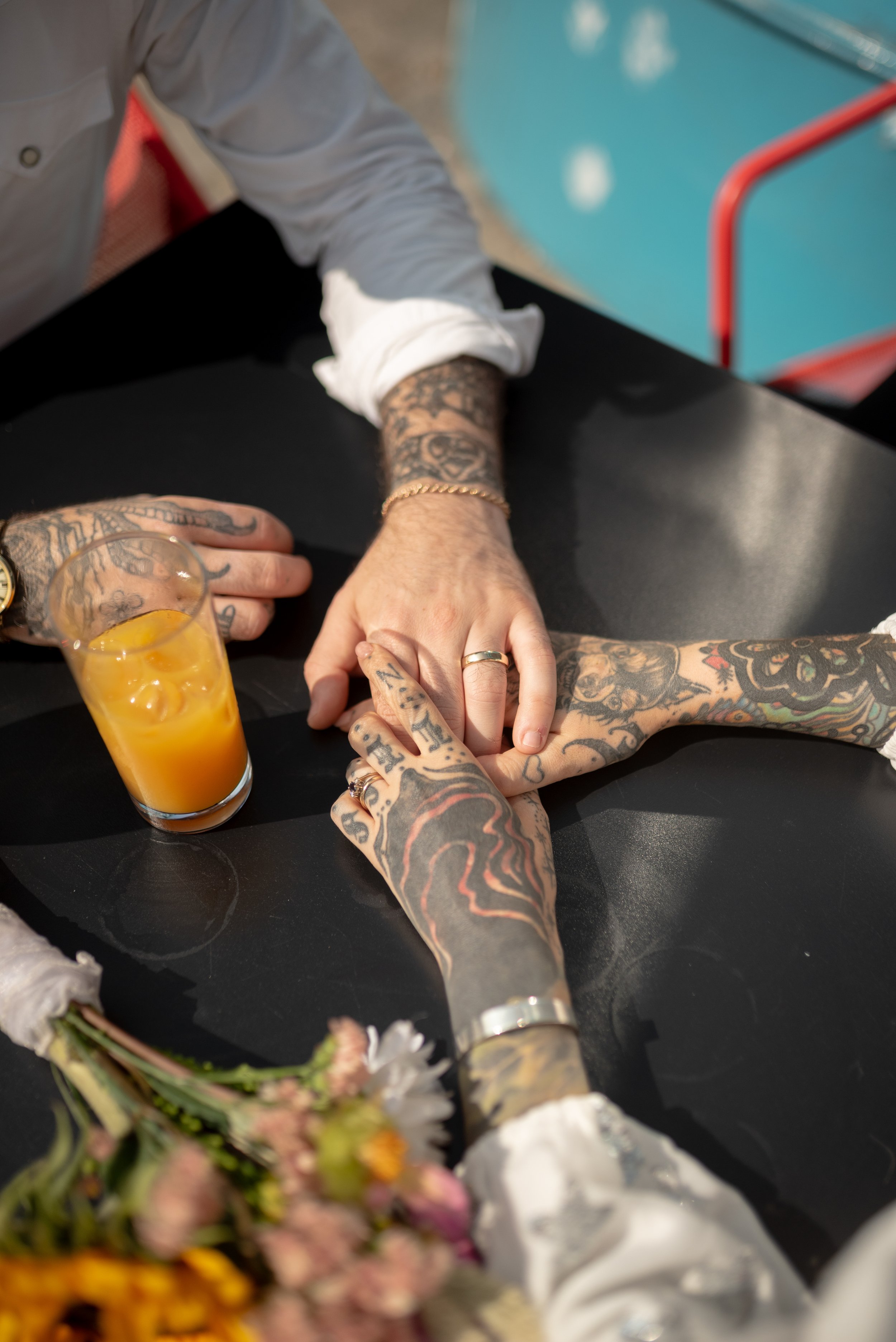 Three tattooed hands, one with a wedding band, touching on a black table near a glass of yellow-orange juice and a bouquet of mixed flowers in the foreground.