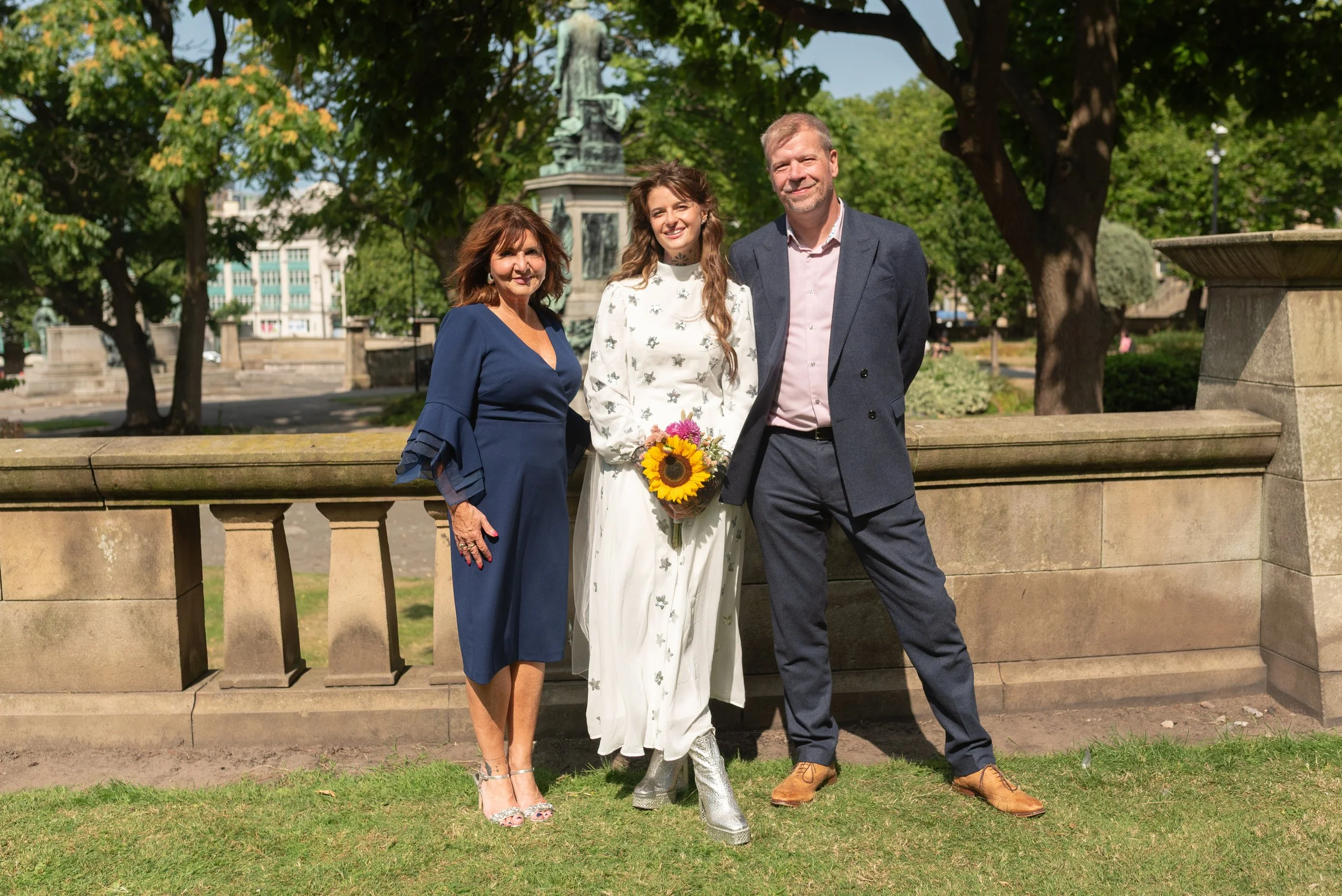 Three people standing together outdoors on a sunny day, two women and one man, with trees and a monument in the background. The woman in the middle is holding a sunflower bouquet and wearing a white dress, the woman on the left is wearing a blue dres