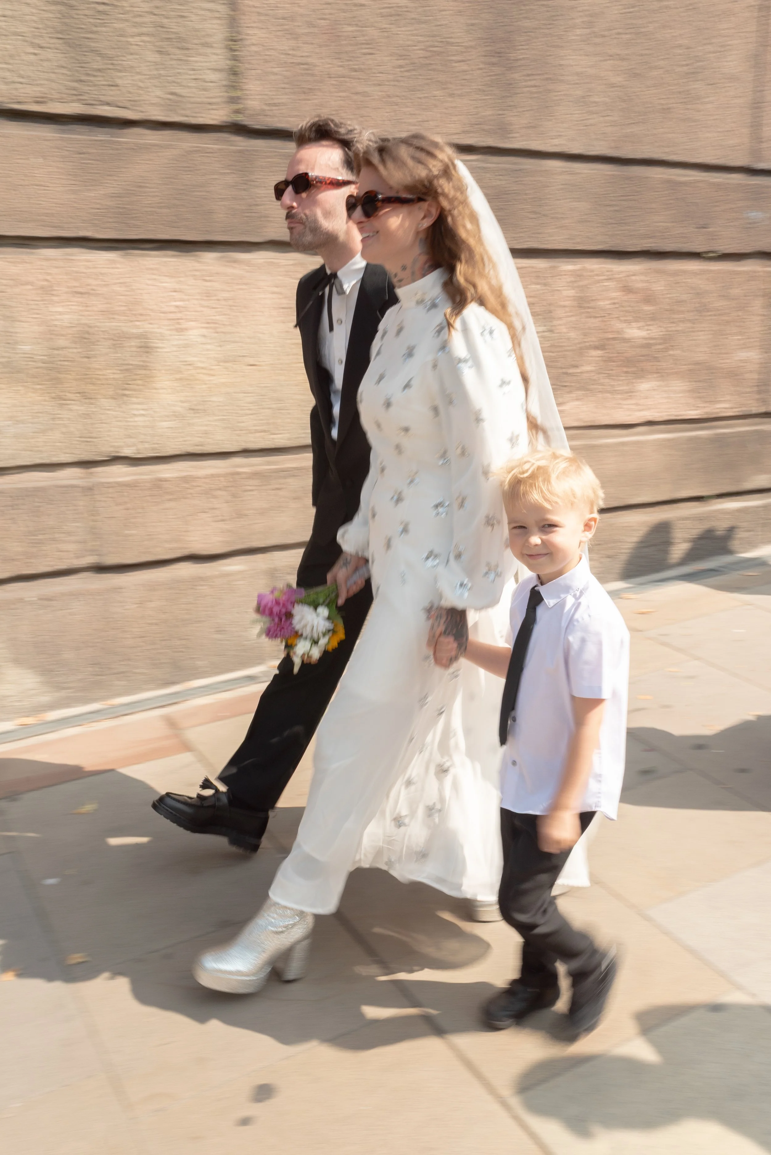 A family walking hand in hand on the sidewalk. The woman is wearing a white floral dress, white platform boots, and sunglasses, holding a small bouquet of flowers. The man is dressed in a black suit with sunglasses. The young boy is wearing a white s