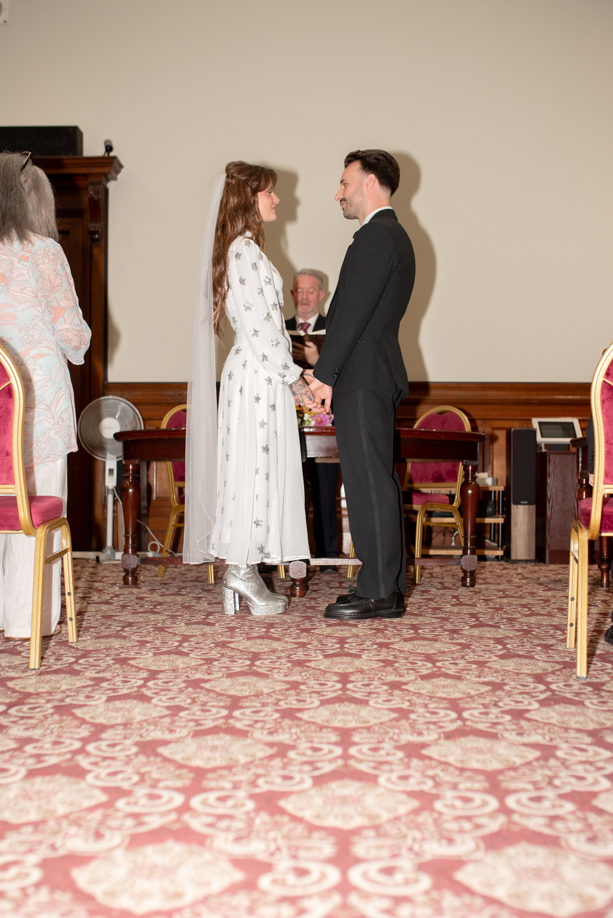 A bride and groom holding hands during a wedding ceremony, facing each other and smiling, in a room with a patterned carpet and red chairs.