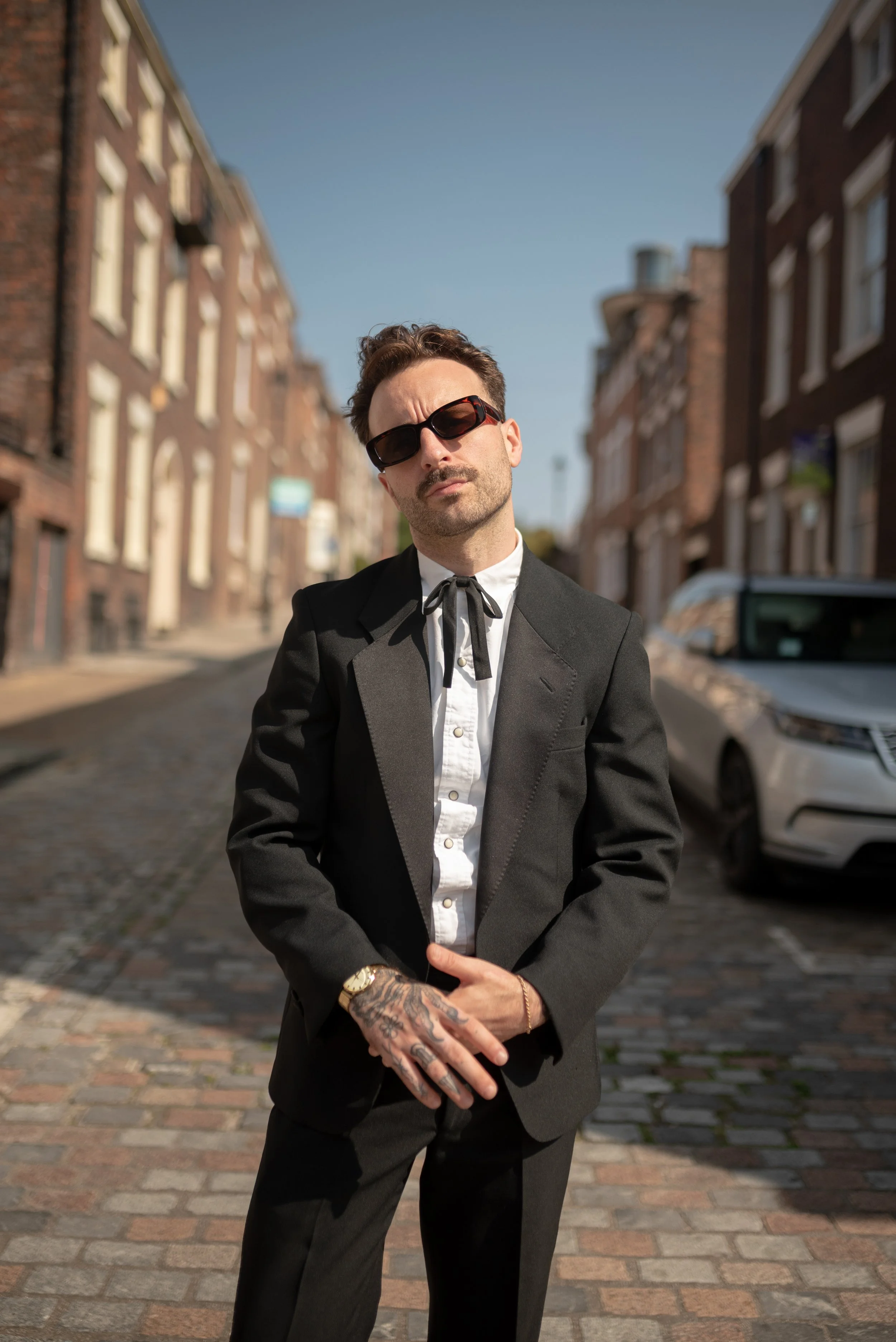 A man in a black suit with a white shirt, black ribbon tie, and sunglasses standing on a cobblestone street with brick buildings and a car in the background.