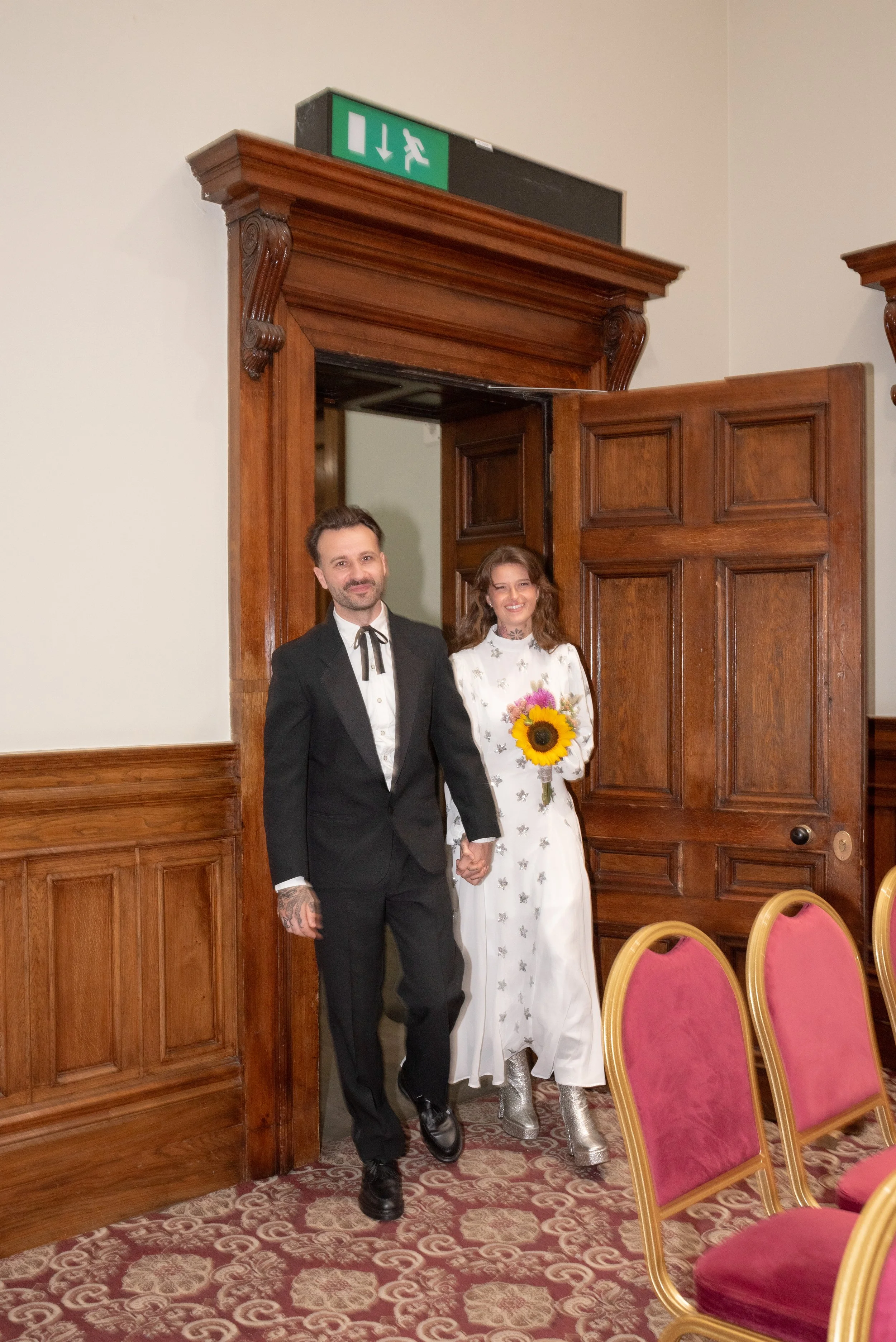 A couple, dressed in formal attire, holding hands and walking out of a room with ornate wooden doors. The woman is holding a sunflower bouquet and wearing silver boots, while the man is wearing a black tuxedo with a bow tie.