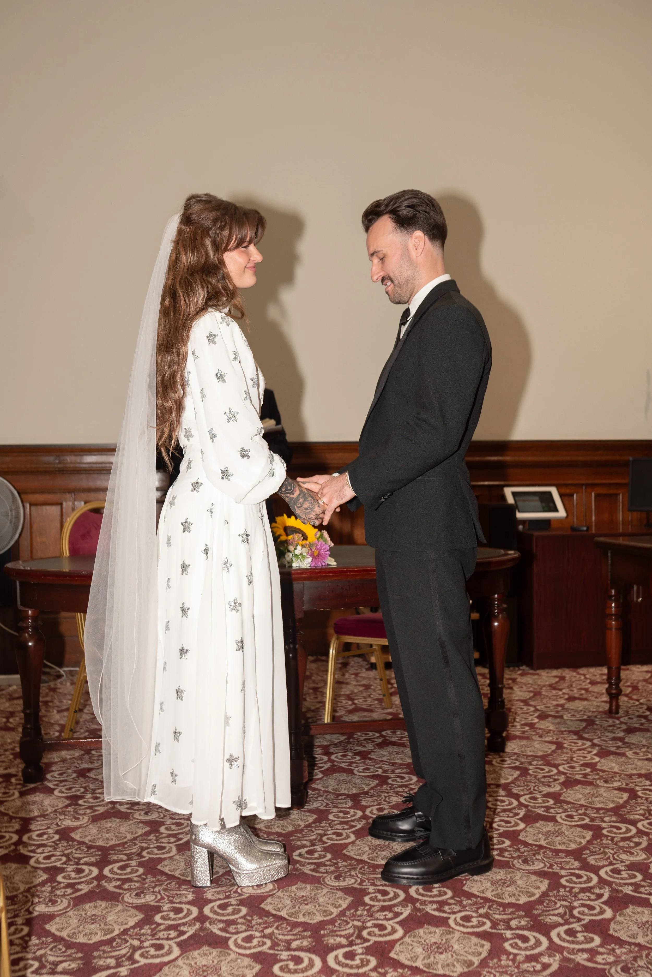 A bride and groom holding hands during their wedding ceremony, standing face to face in a room with wooden paneling, carpeted floor, and chairs.
