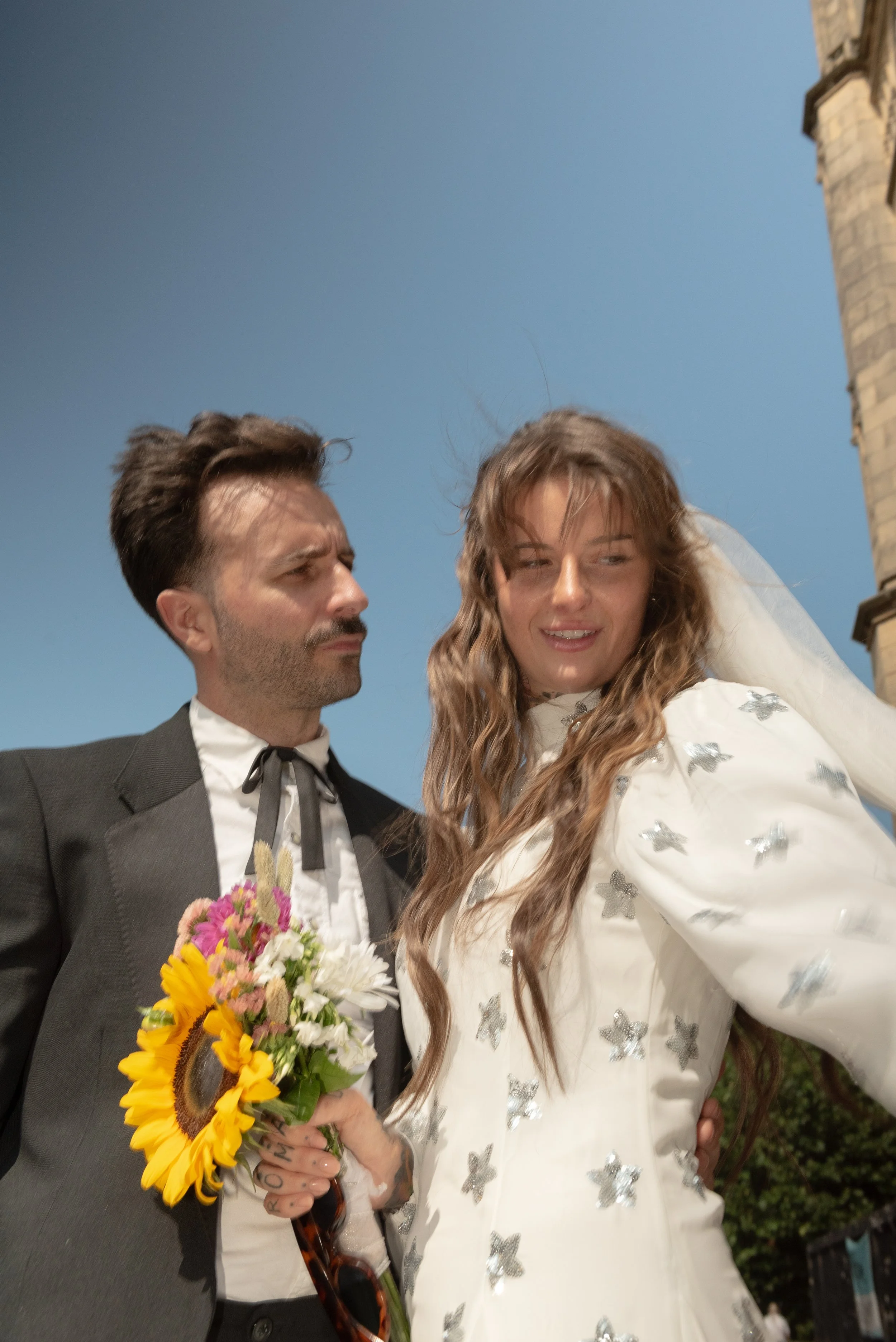 A man holding a bouquet of flowers, wearing a tuxedo, and a woman in a white dress with silver embellishments, possibly a wedding dress, standing outdoors with a brick building and a blue sky in the background.
