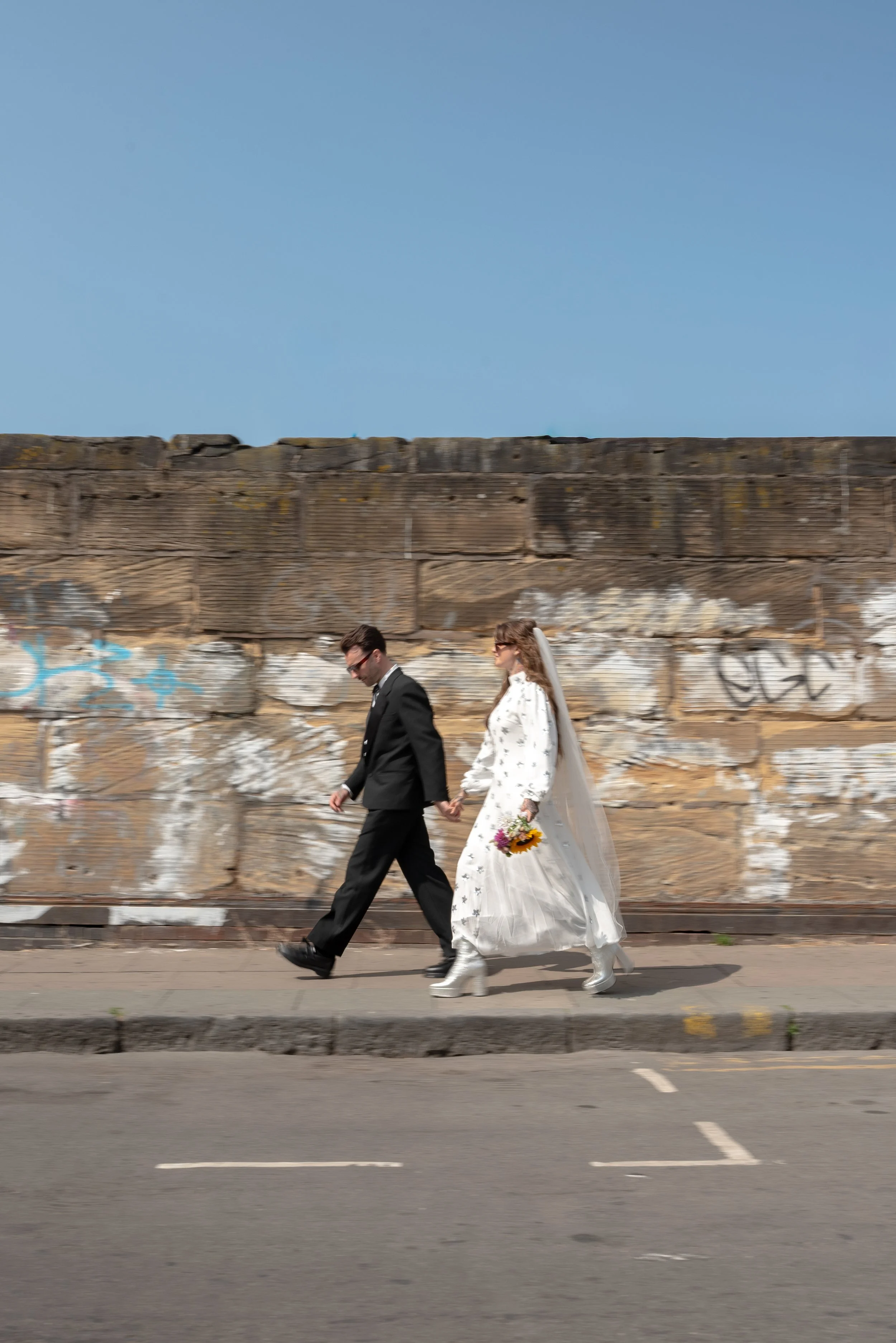 A bride and groom walking hand in hand on the sidewalk, with a graffiti-covered stone wall and a clear blue sky in the background.