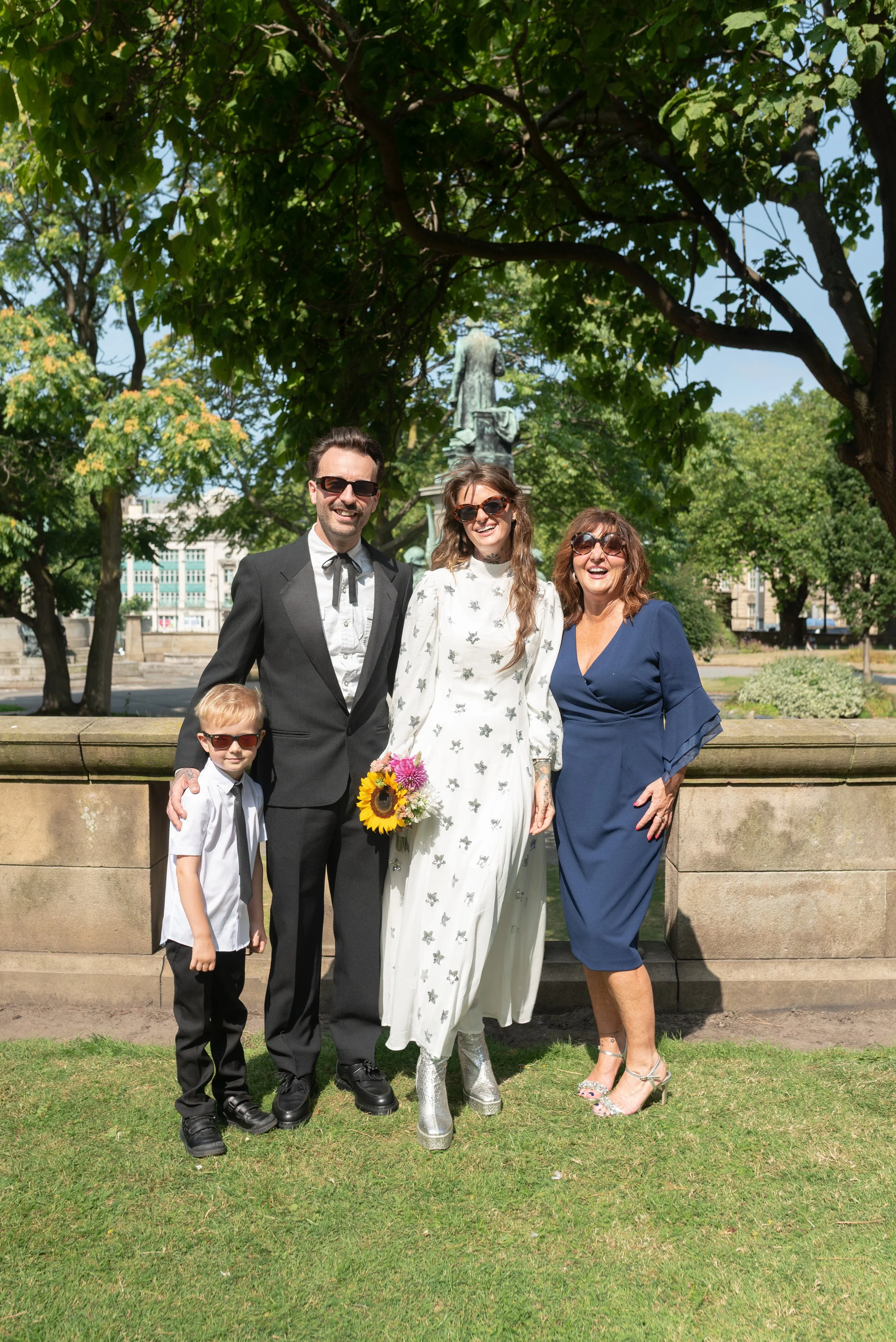 A family of five posing outdoors on a sunny day, standing in front of a stone railing with a statue and trees in the background. The woman in the center is holding a bouquet of sunflowers and wearing a white dress with a floral pattern. The man to he