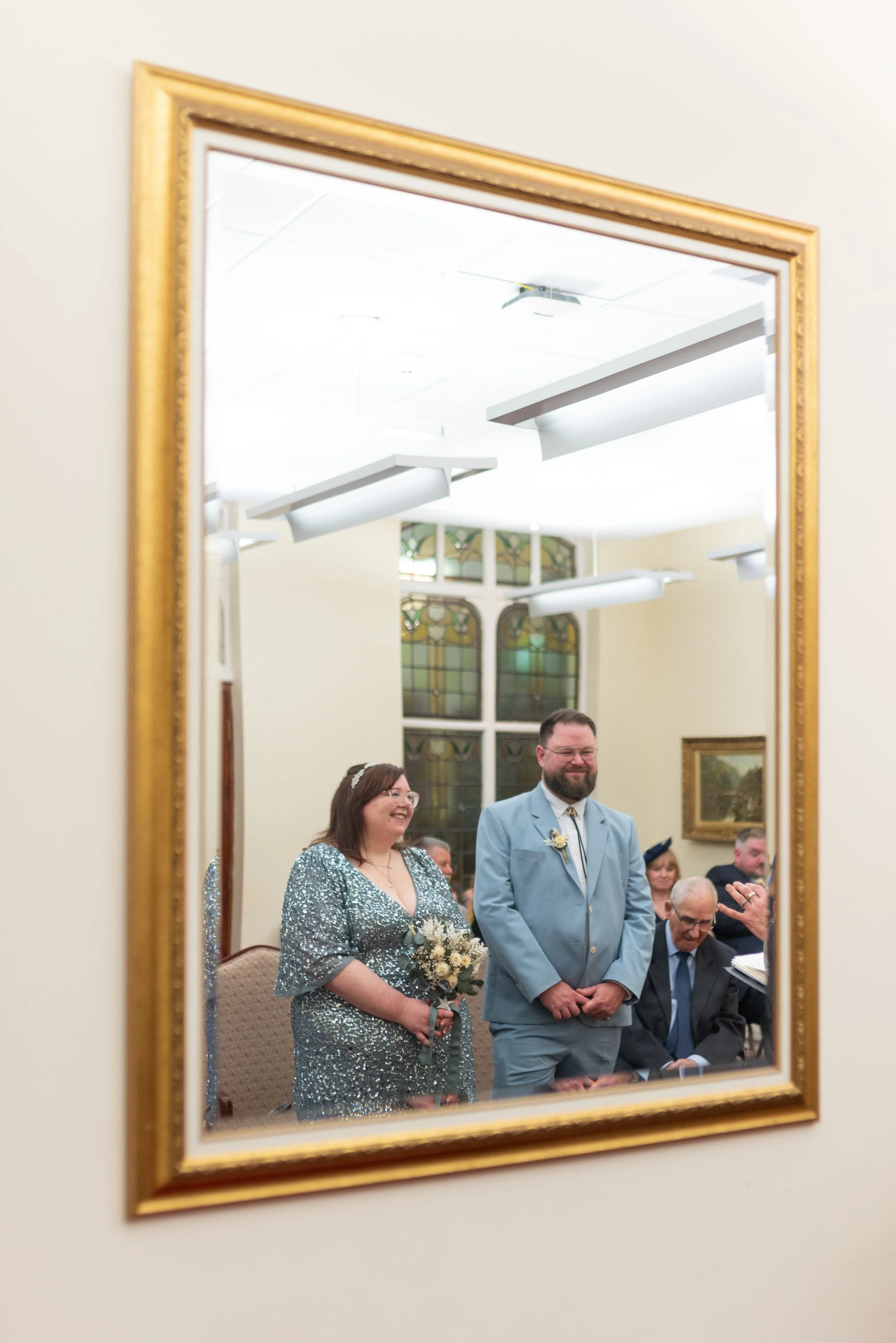 A mirror reflecting a wedding ceremony with a woman in a silver dress holding a bouquet, a man in a light blue suit, and other guests in the background in a church with stained glass windows.