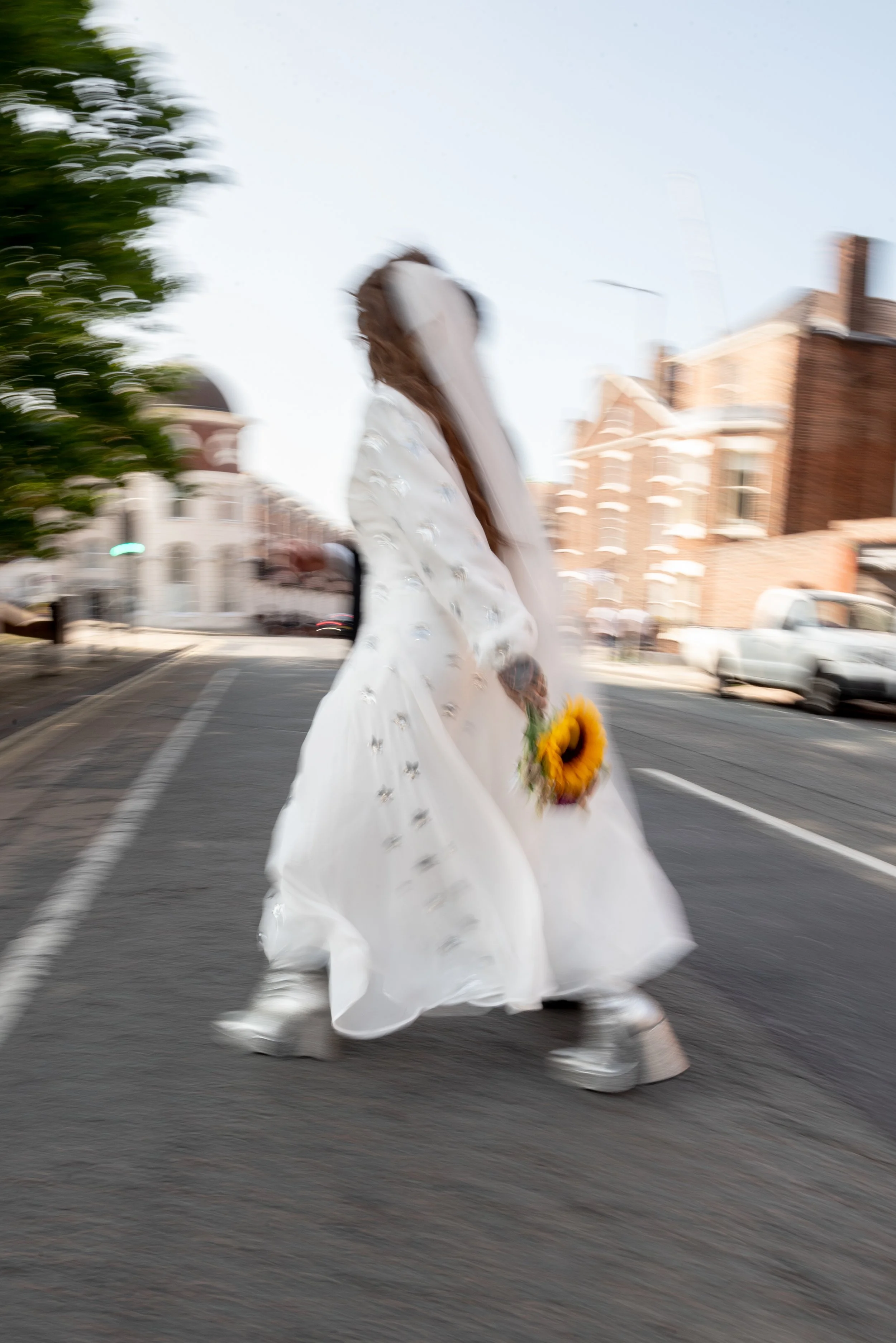 Person in a white dress walking across the street, holding a sunflower, with a city street background and motion blur.