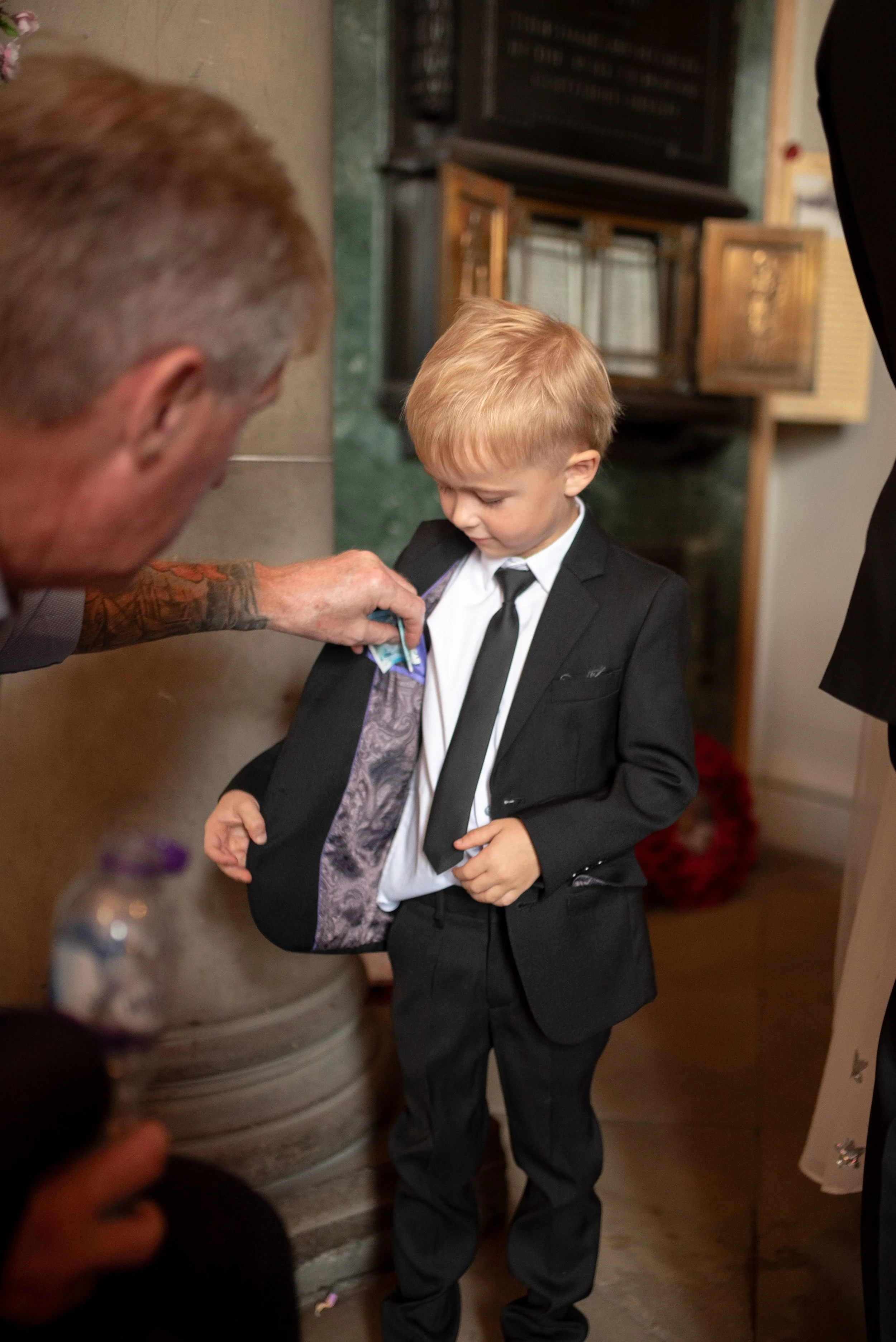 A young boy in a black suit and tie looks down at a pocket square being placed in his jacket by an adult with tattoos on his arm. The scene appears to be inside a decorated indoor space, possibly during a formal event.
