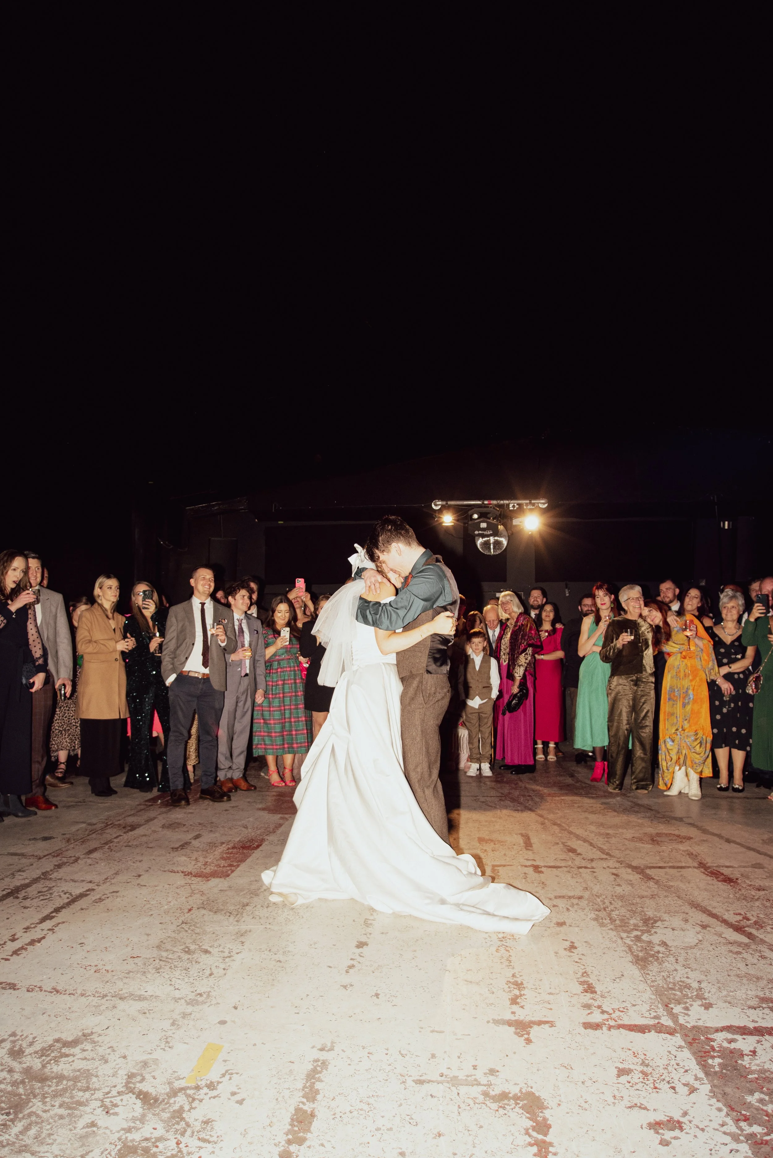 A bride and groom dancing at their wedding reception, surrounded by guests watching and taking photos indoors at night.