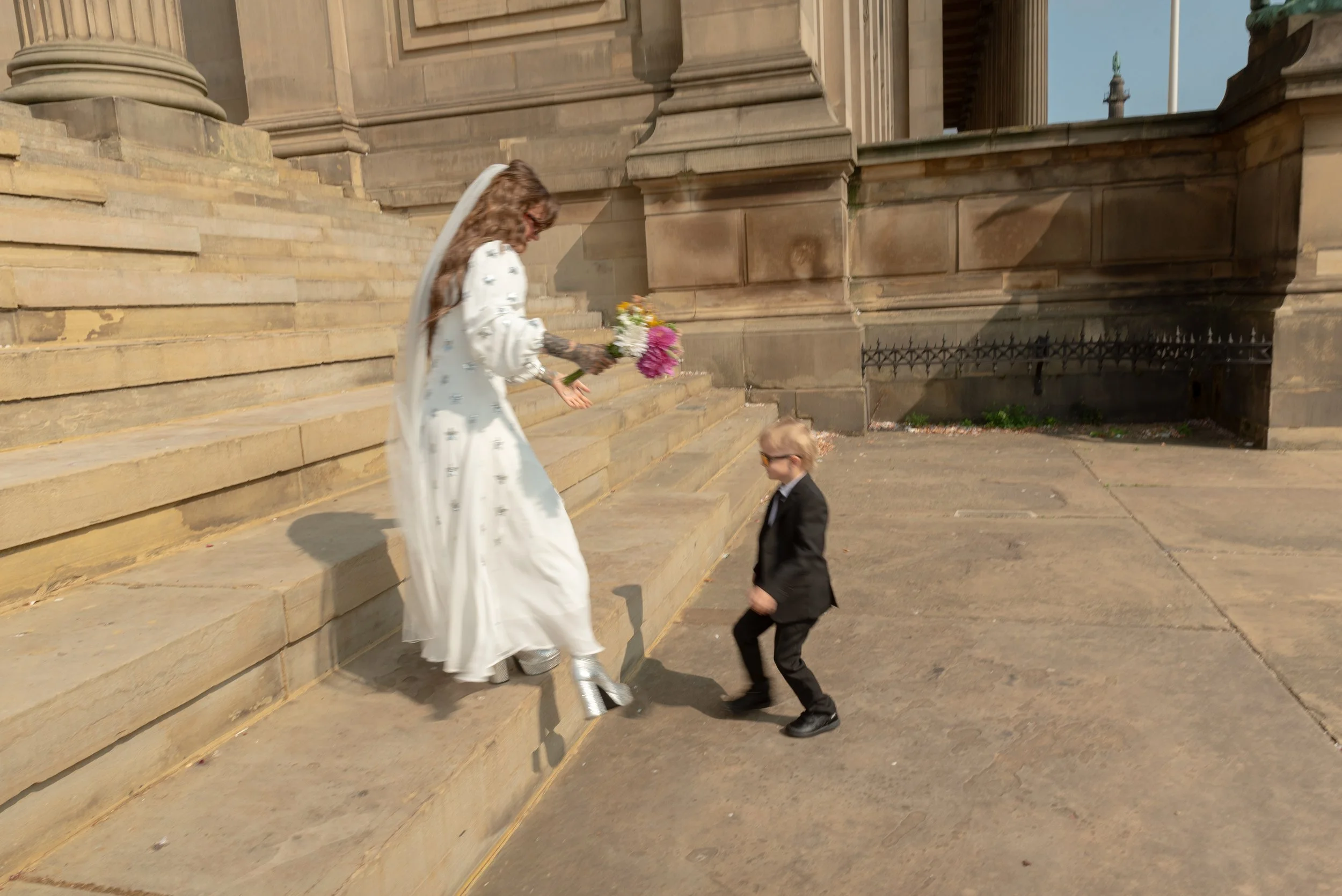A woman in a white dress with a long veil, holding a bouquet of flowers, descending stone steps outside a large historic building while a young boy in a black suit and sunglasses looks up at her.