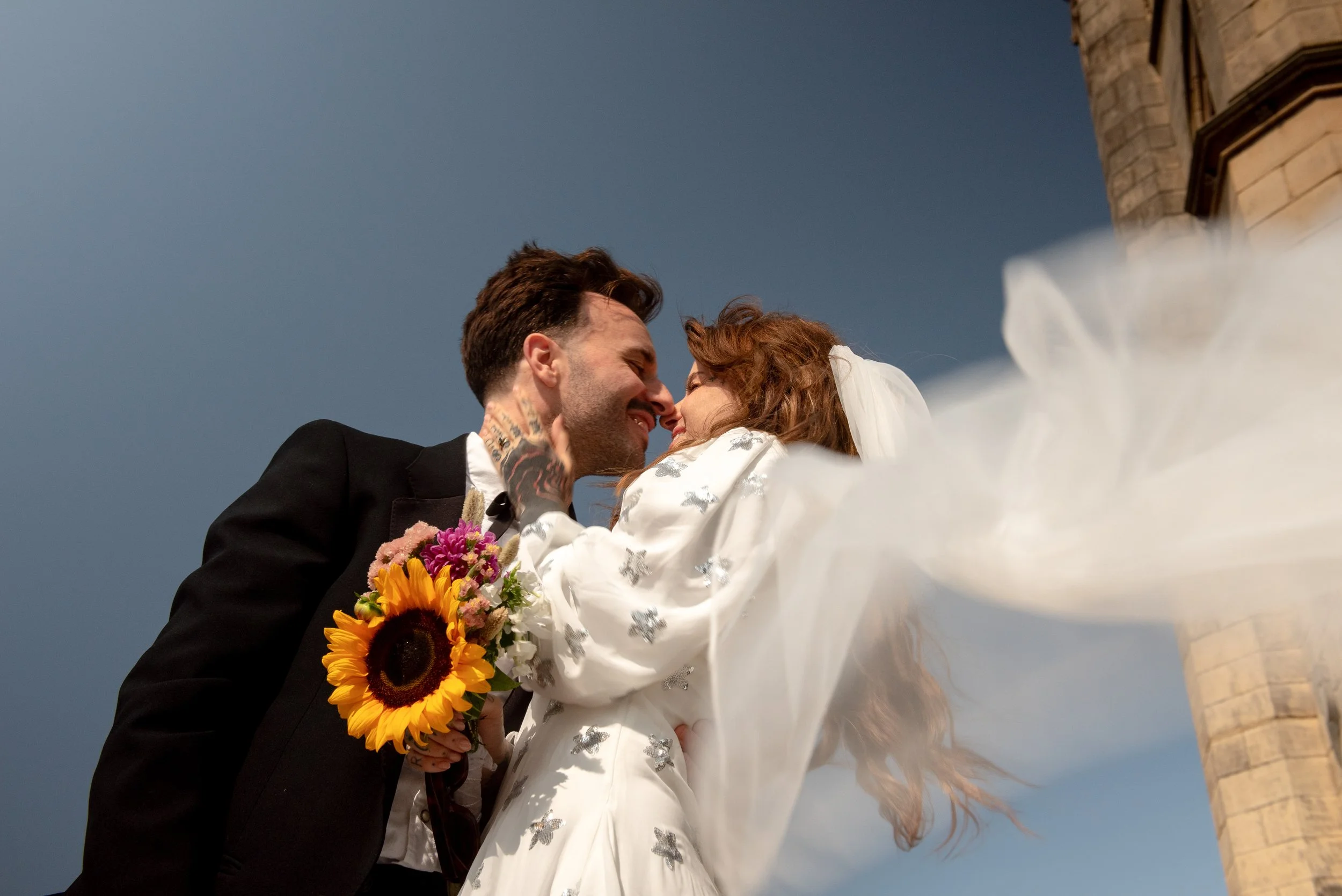 A couple in wedding attire sharing a kiss outdoors, with the bride holding a bouquet of sunflowers and other flowers, and a brick tower in the background against a cloudy sky.