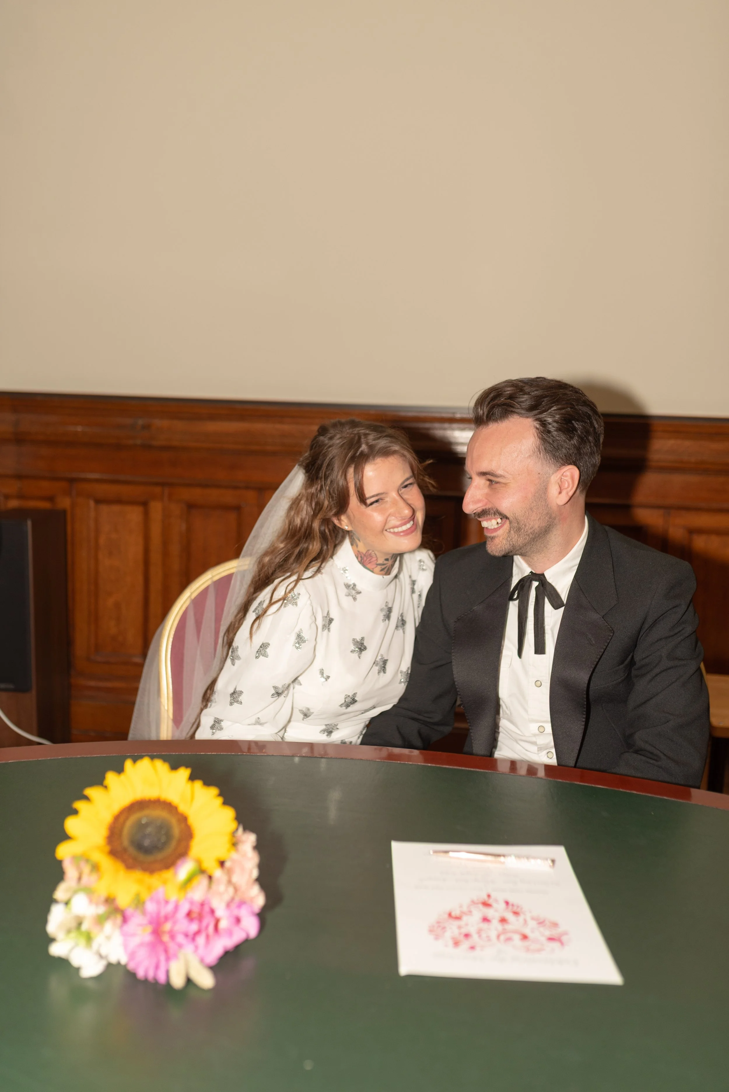 A bride and groom sitting at a table, smiling and laughing together during their wedding. The bride has brown hair, a white dress with floral details, and a veil, while the groom has dark hair and a black tuxedo with a bowtie. There is a bouquet of s