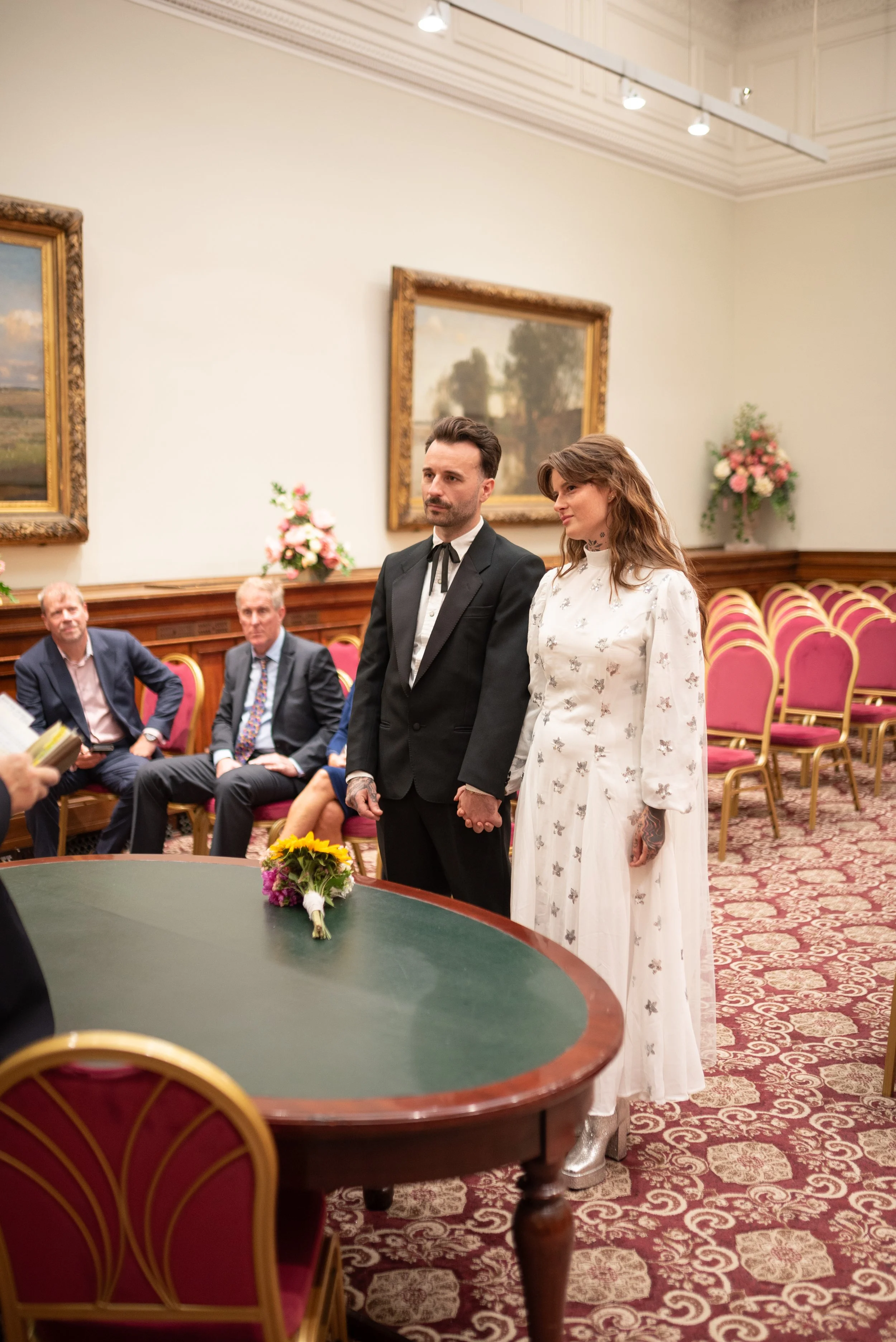 A couple getting married, holding hands and standing in a formal room with pink chairs, framed paintings, and floral arrangements, during a wedding ceremony.