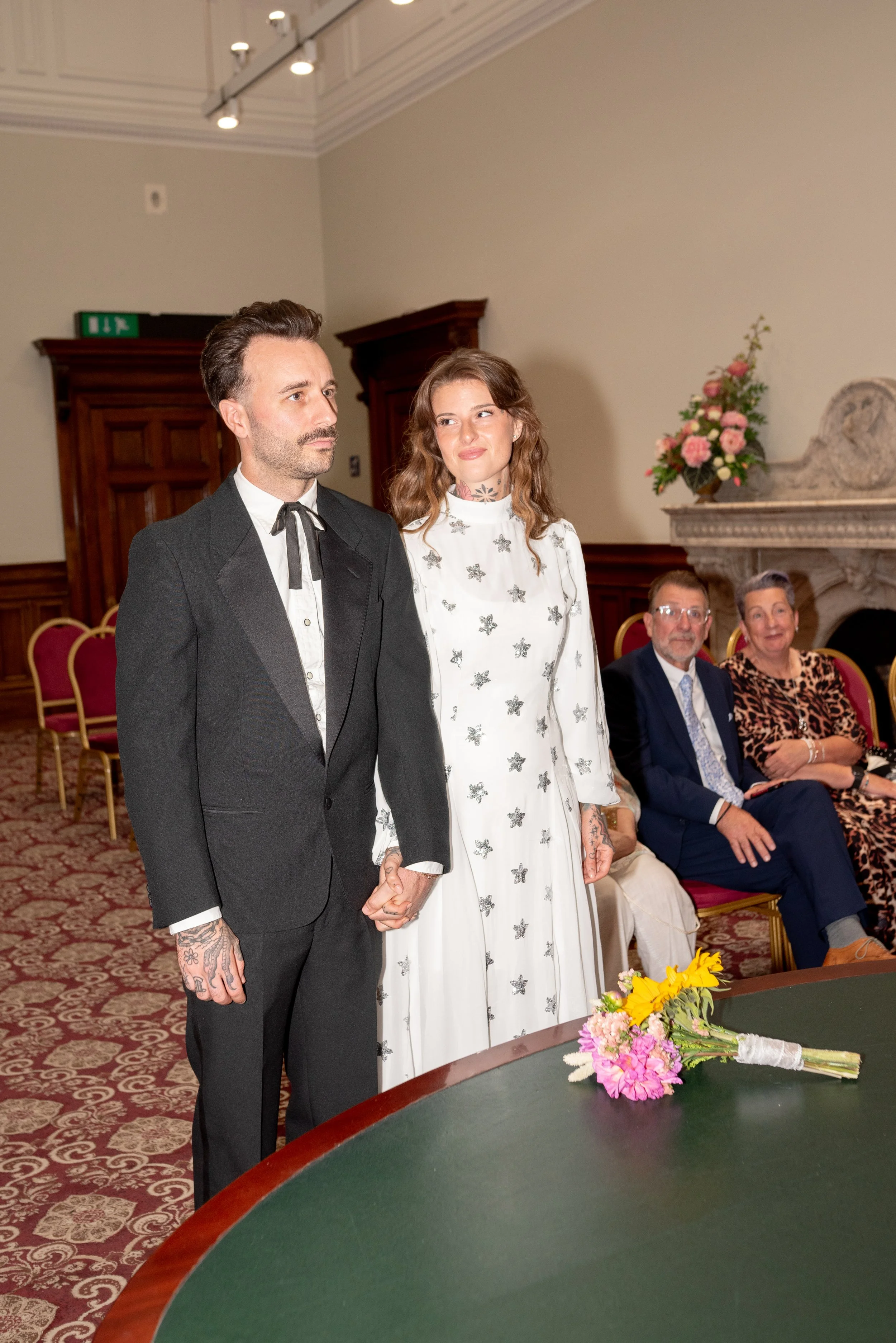 A couple holding hands during a wedding ceremony, with four seated guests in the background, in a room with wood-paneled walls and a fireplace, flowers on a table in the foreground.