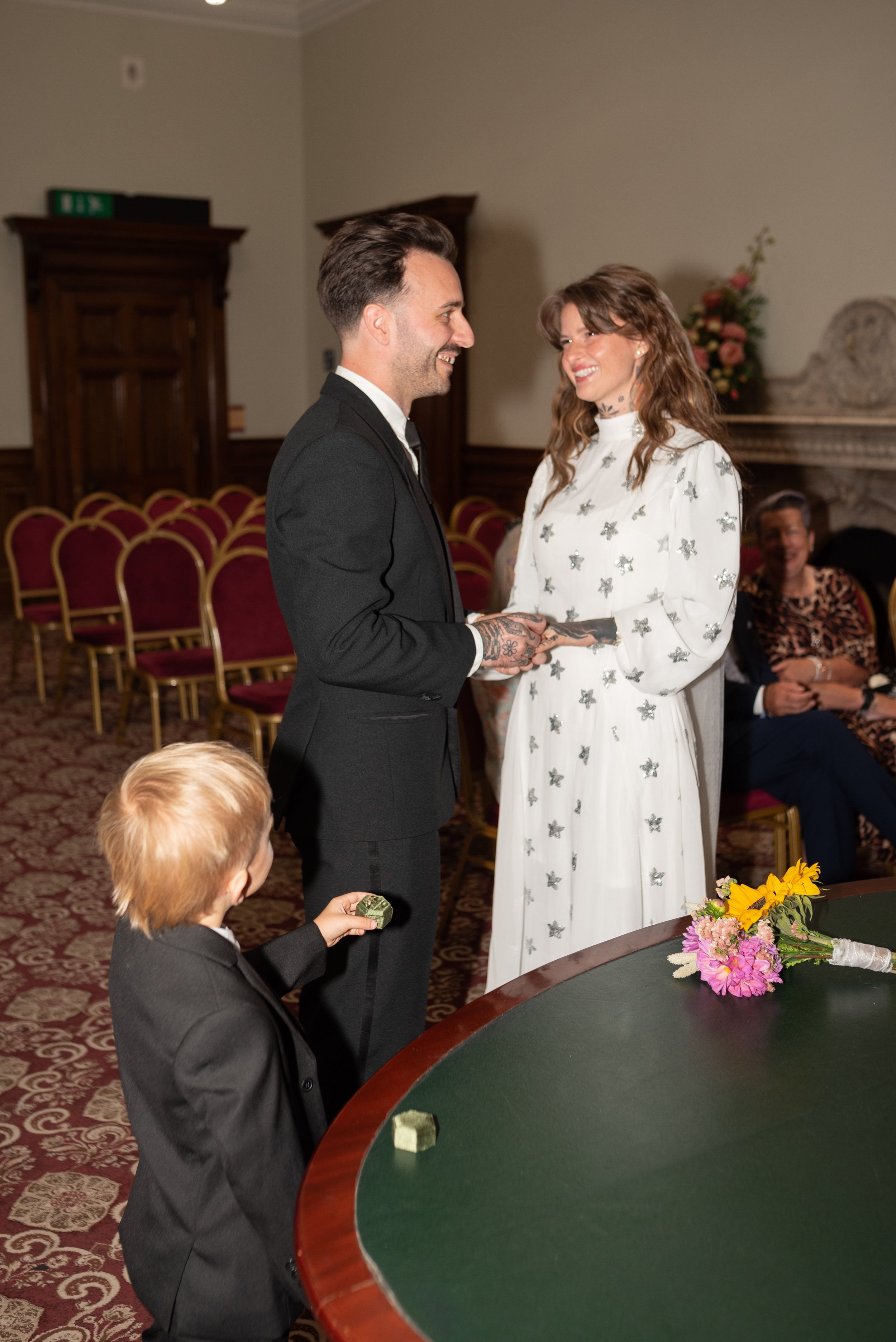 A woman in a white dress with silver star patterns and long sleeves exchanges vows with a man in a black suit during a wedding ceremony. The woman is smiling at the man, who is smiling back. A small child with blond hair in a black suit holds a small