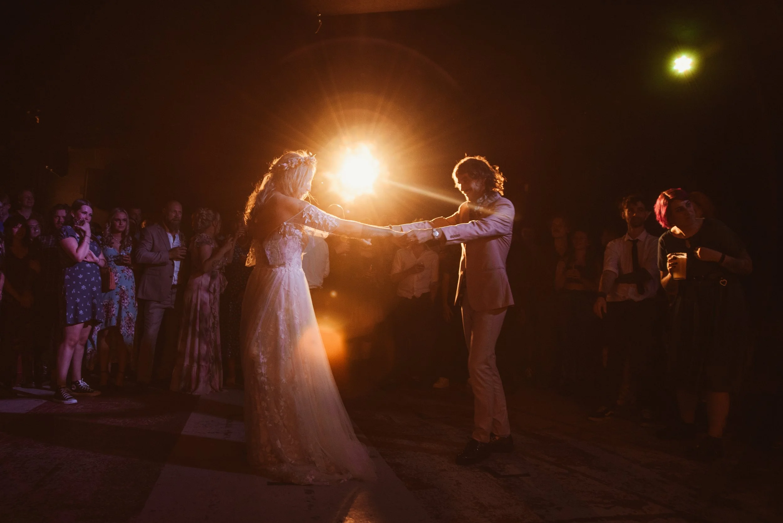 A bride and groom dancing at their wedding reception during sunset, surrounded by guests in a dimly lit venue with warm lighting.