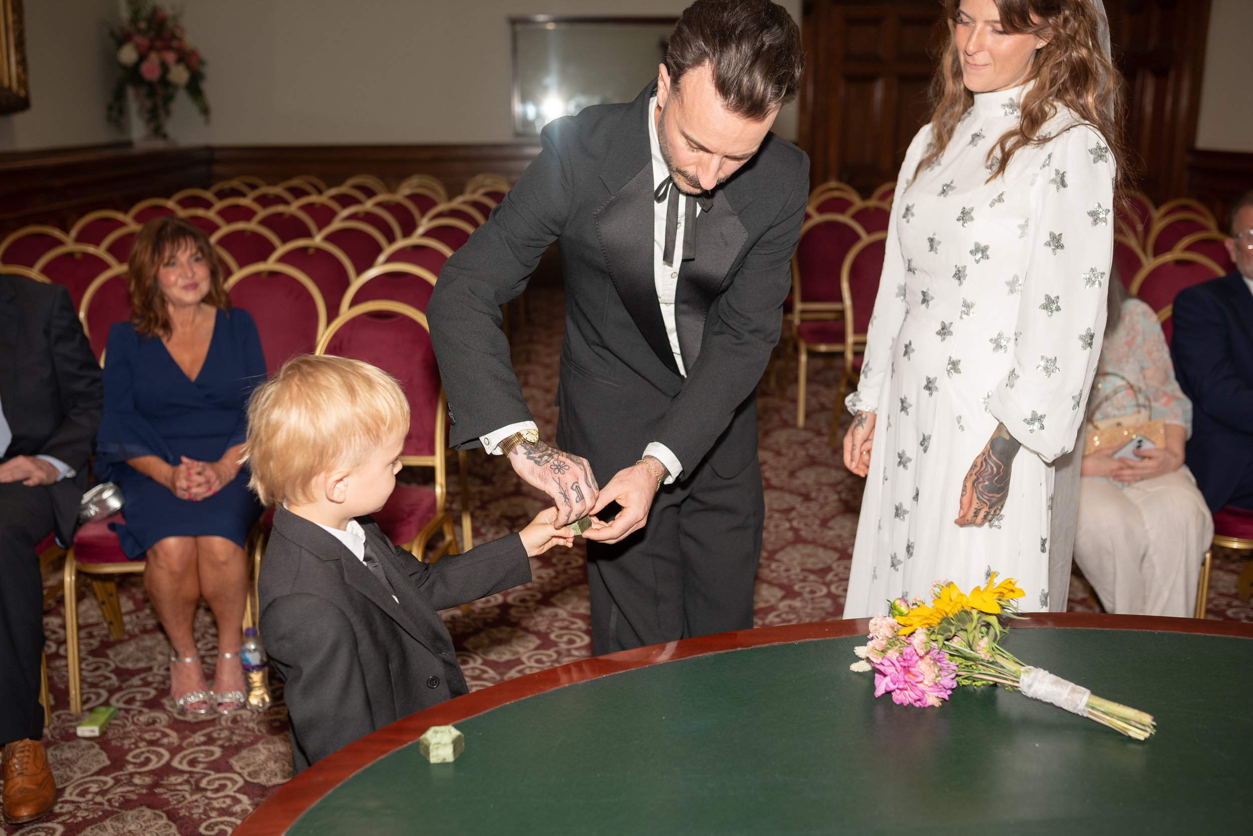 A wedding ceremony where a man in a suit places a ring on a young boy's finger. A woman in a white dress with butterfly patterns watches. There are seated guests, including a woman in a blue dress, and a bouquet of flowers on a table.