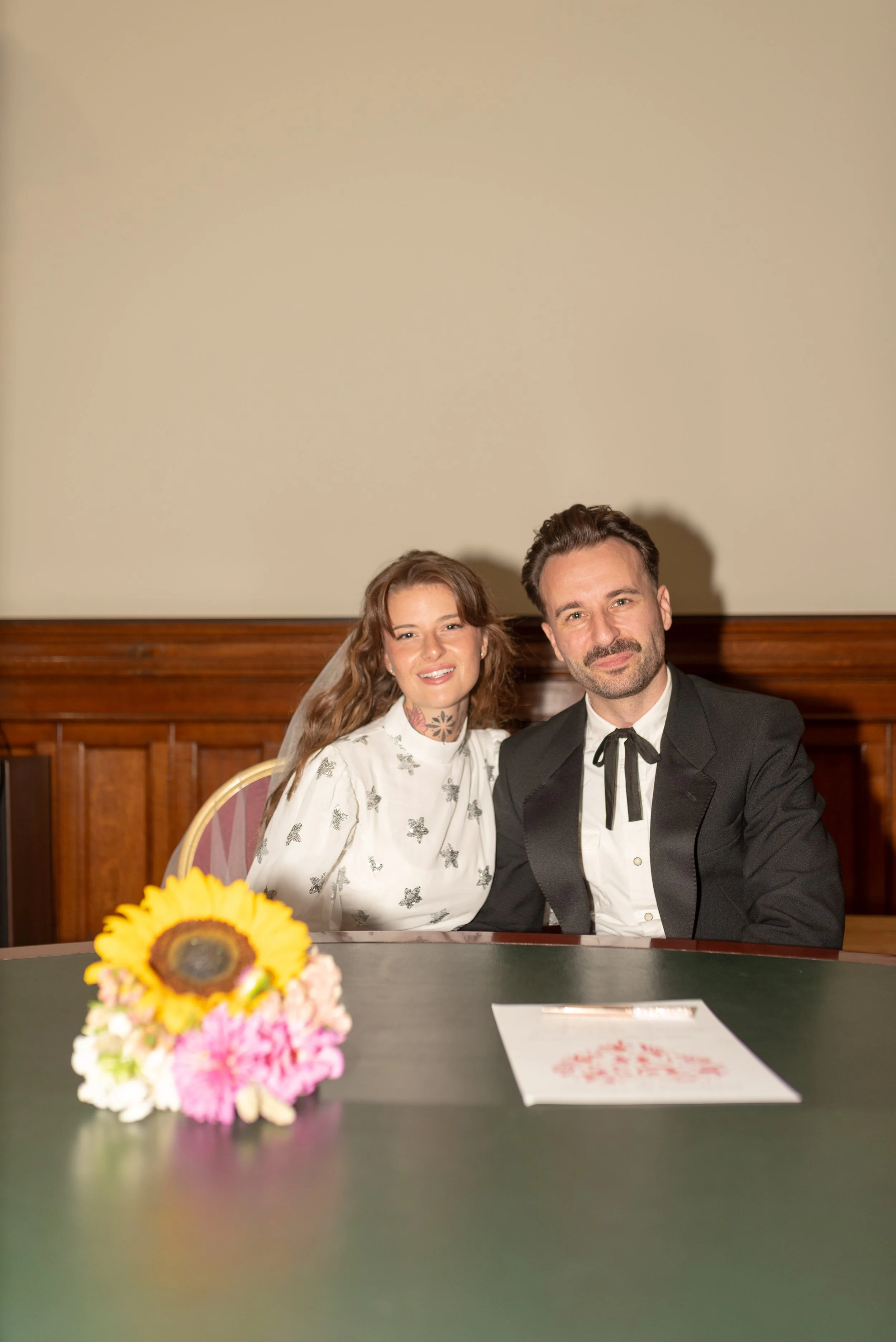 A couple sitting together at a table with a bouquet of sunflowers and pink flowers, and a document, in a room with wooden paneling.