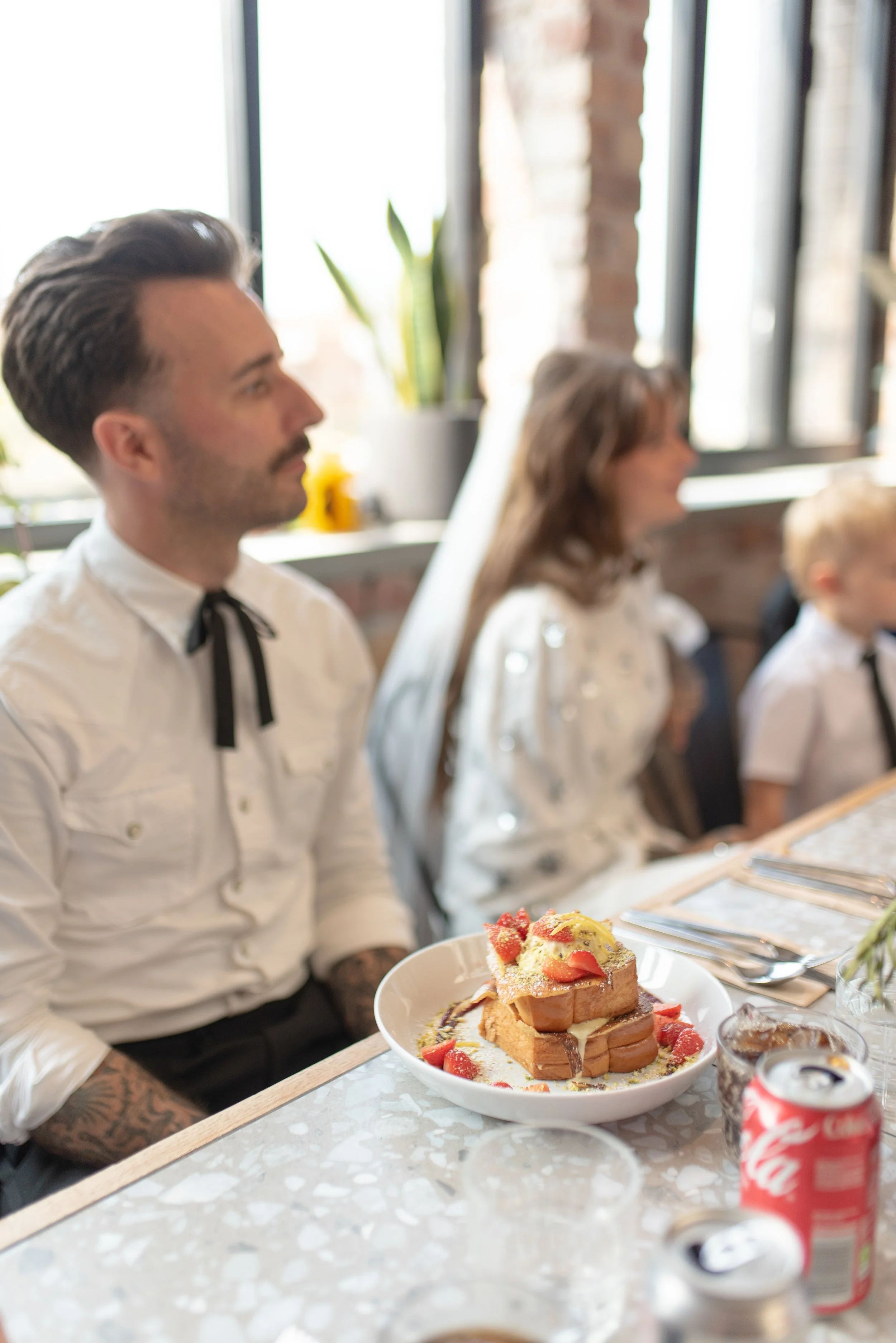 A man with dark hair and tattoos on his arms is sitting at a table in a bright room, alongside a woman and a child. The table has a plate with stacked bread and strawberries, and cans of soda.