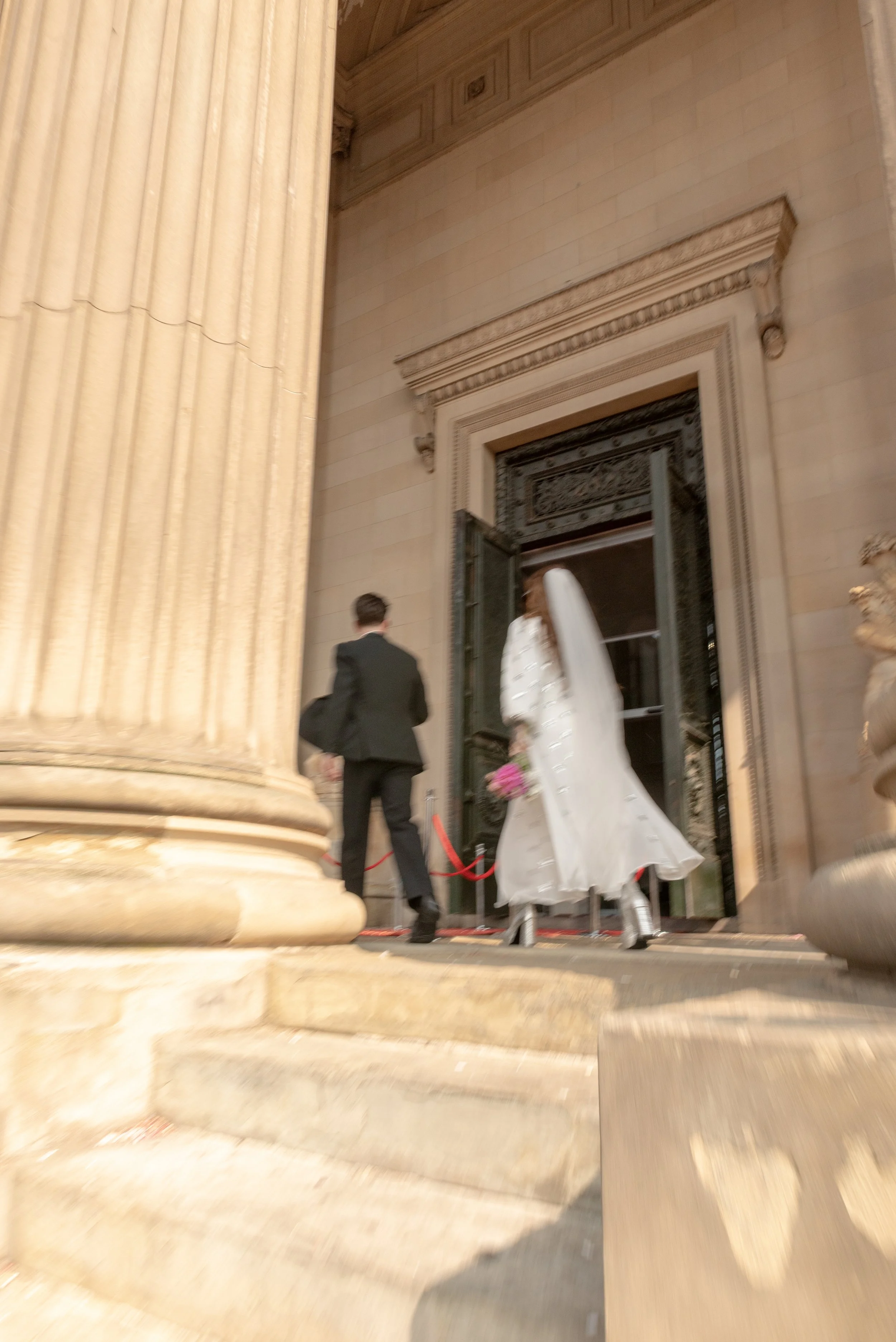 A bride in a white wedding dress and veil holding a bouquet of pink flowers and a groom in a black suit walking up steps outside a grand building.