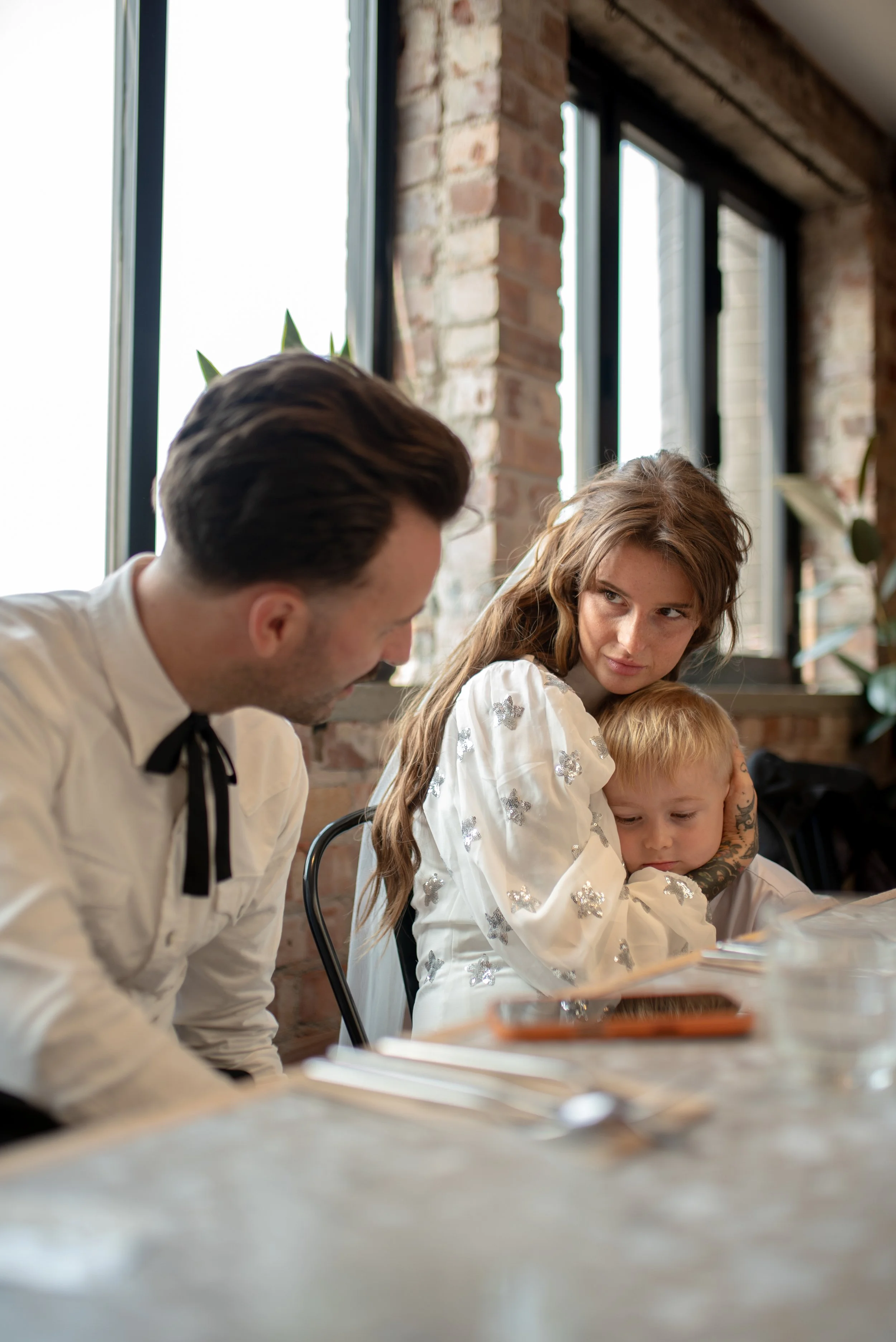 A woman holds a young child on her lap while a man leans in, all sitting at a table in a restaurant with brick walls and large windows.