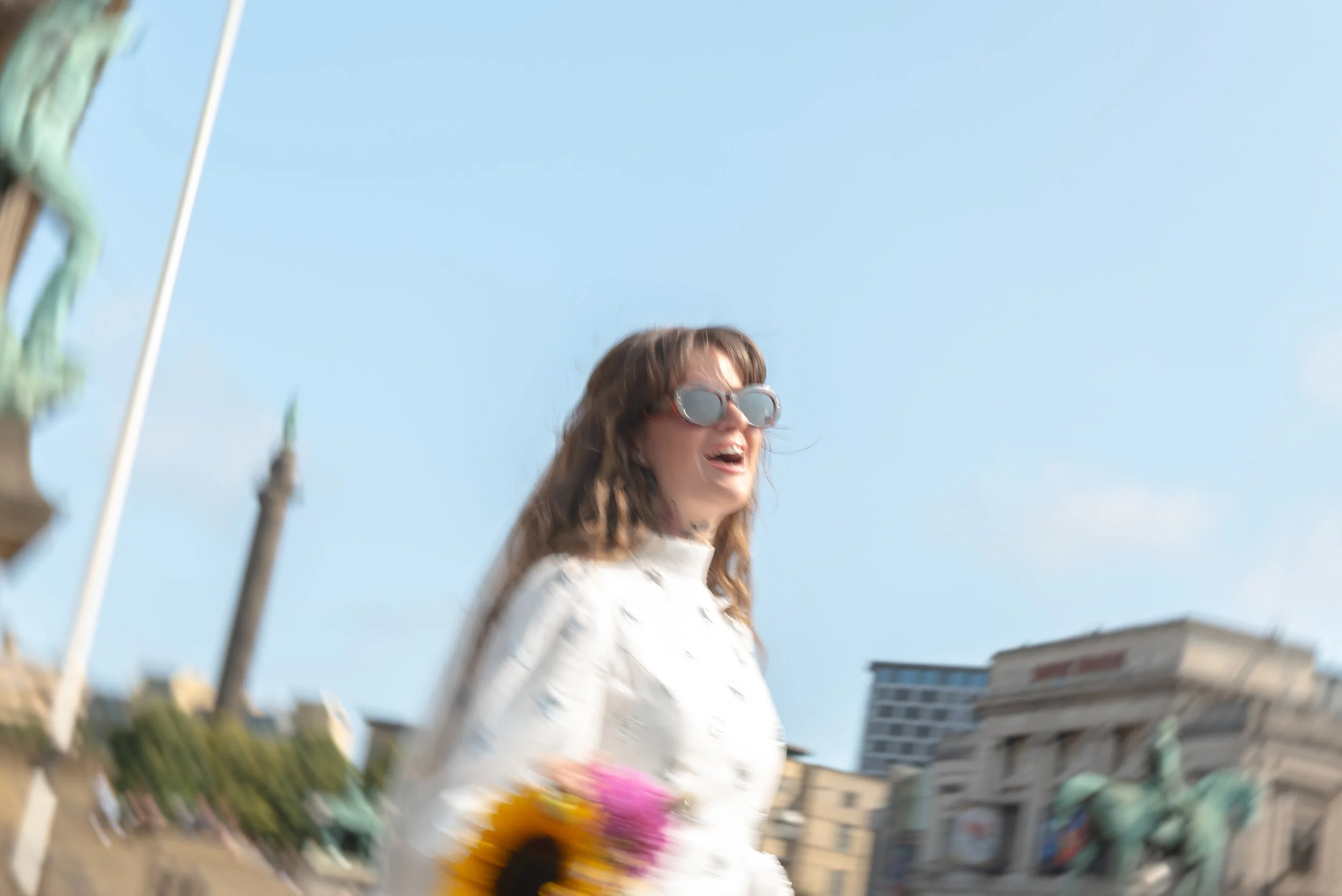 Blurred image of a woman with wavy brown hair wearing large sunglasses and a white jacket, holding a bunch of colorful flowers, standing outdoors in front of a building and statue on a sunny day.