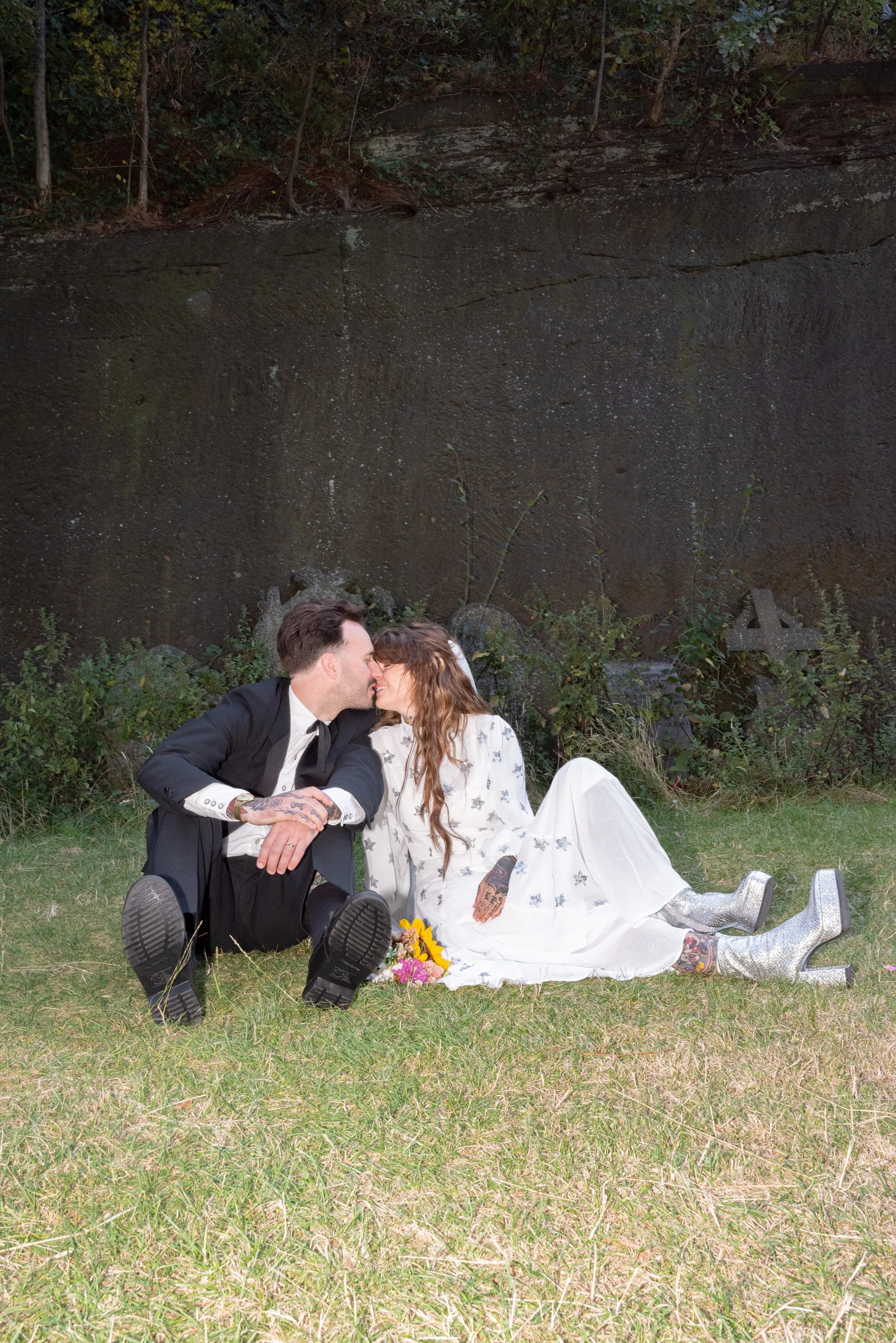 A couple dressed in formal attire sitting on the grass, about to kiss, with a floral bouquet nearby, in front of a dark, moss-covered wall with some greenery and a cross-shaped object.