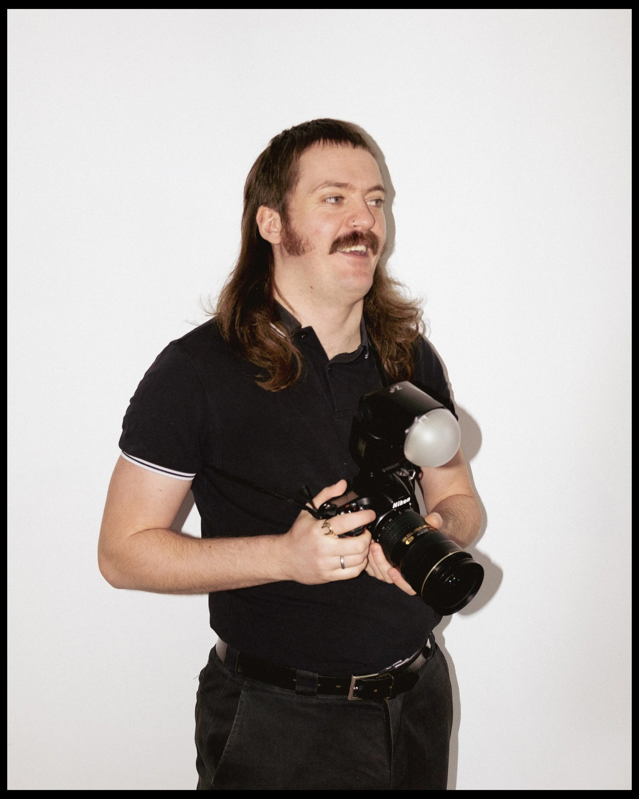 A man with long brown hair, a mustache, and beard holding a professional camera, standing against a plain white background.