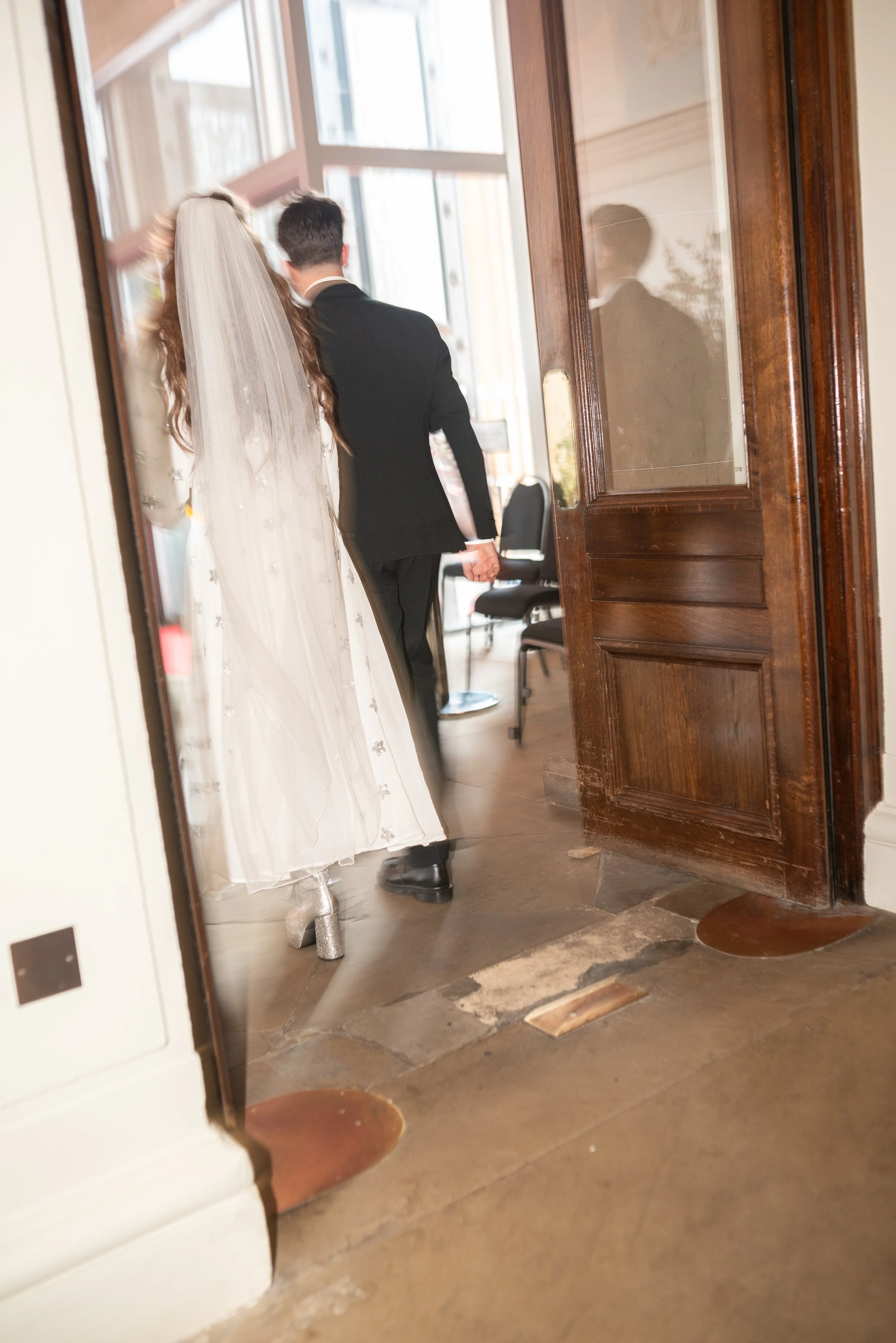 A bride and groom walking through a doorway into a venue, seen through a mirror reflection. The bride wears a white wedding dress and veil, and the groom wears a black suit.