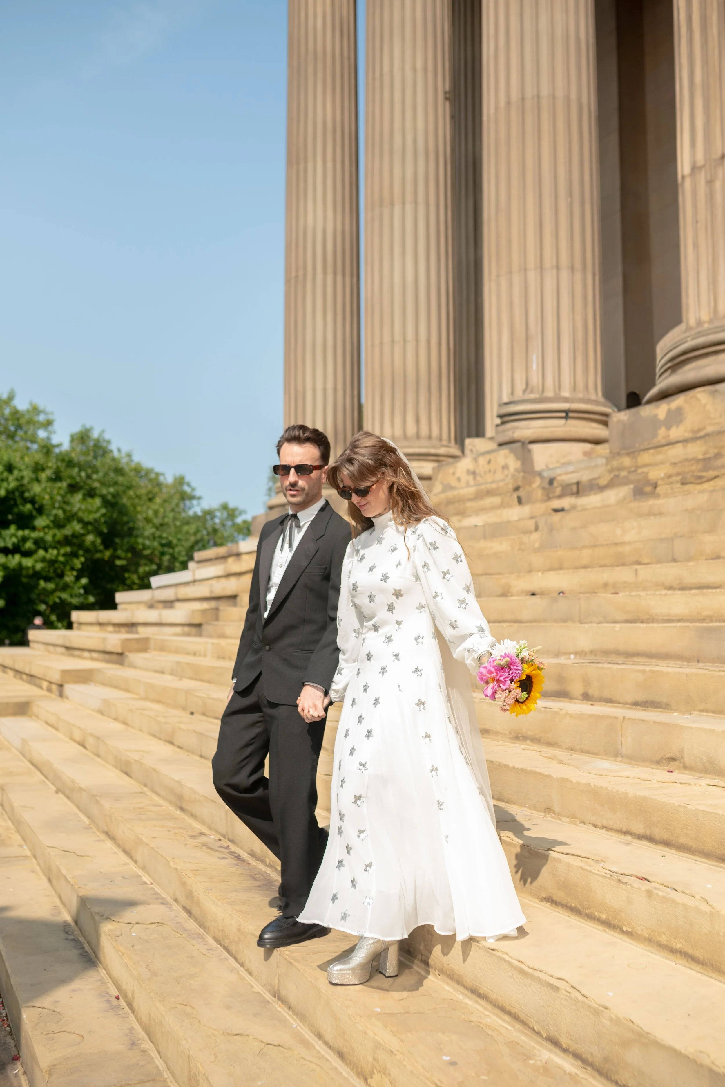 A couple, dressed in wedding attire, walking down the steps of a historic building with large columns. The bride is holding a bouquet of flowers and wearing a white dress with silver embellishments, and the groom is in a black suit with sunglasses.
