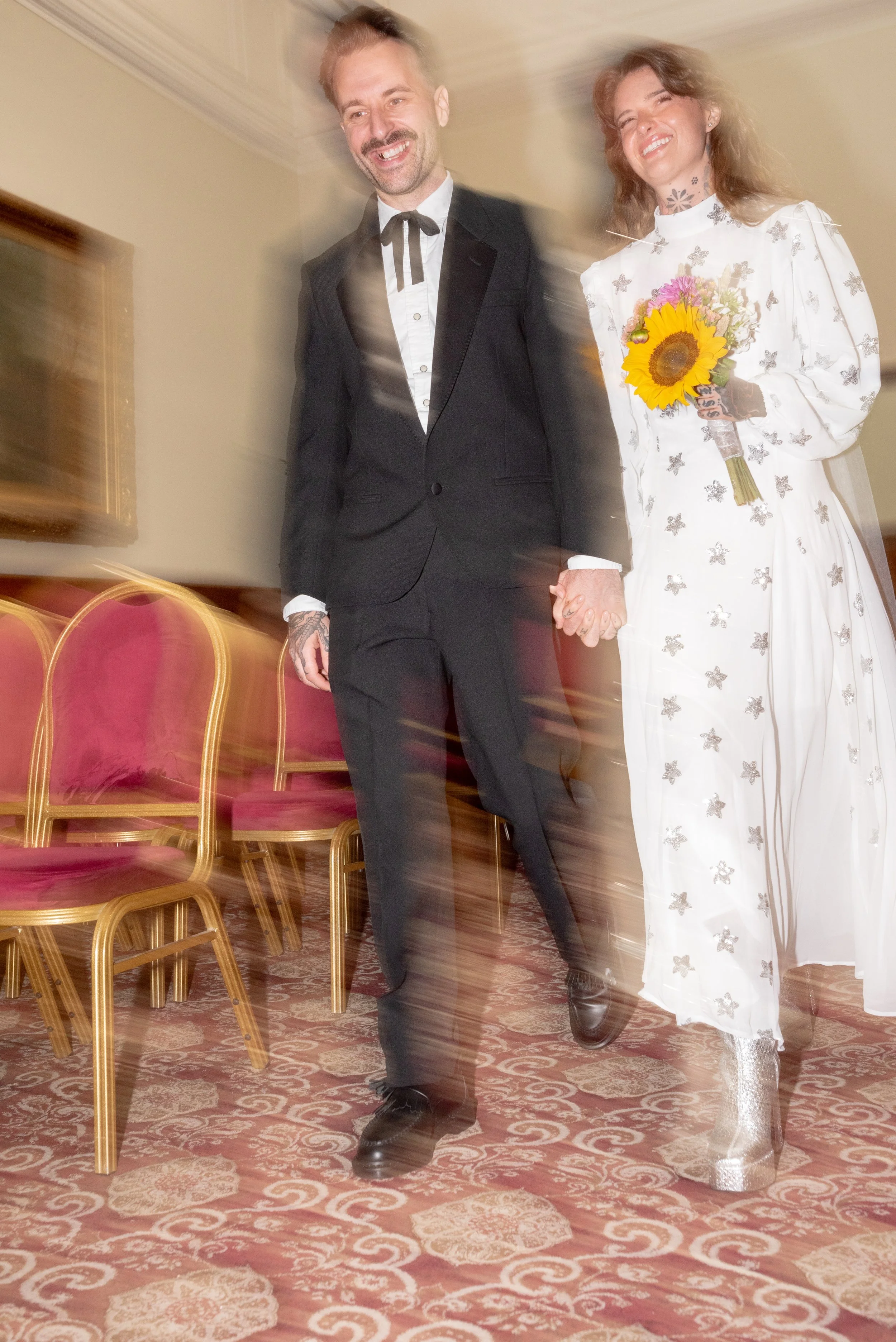 A couple holding hands walking down a room aisle, smiling, dressed in wedding attire; the man in a black suit and bowtie, the woman in a white dress with butterfly print and silver boots, holding a sunflower bouquet.