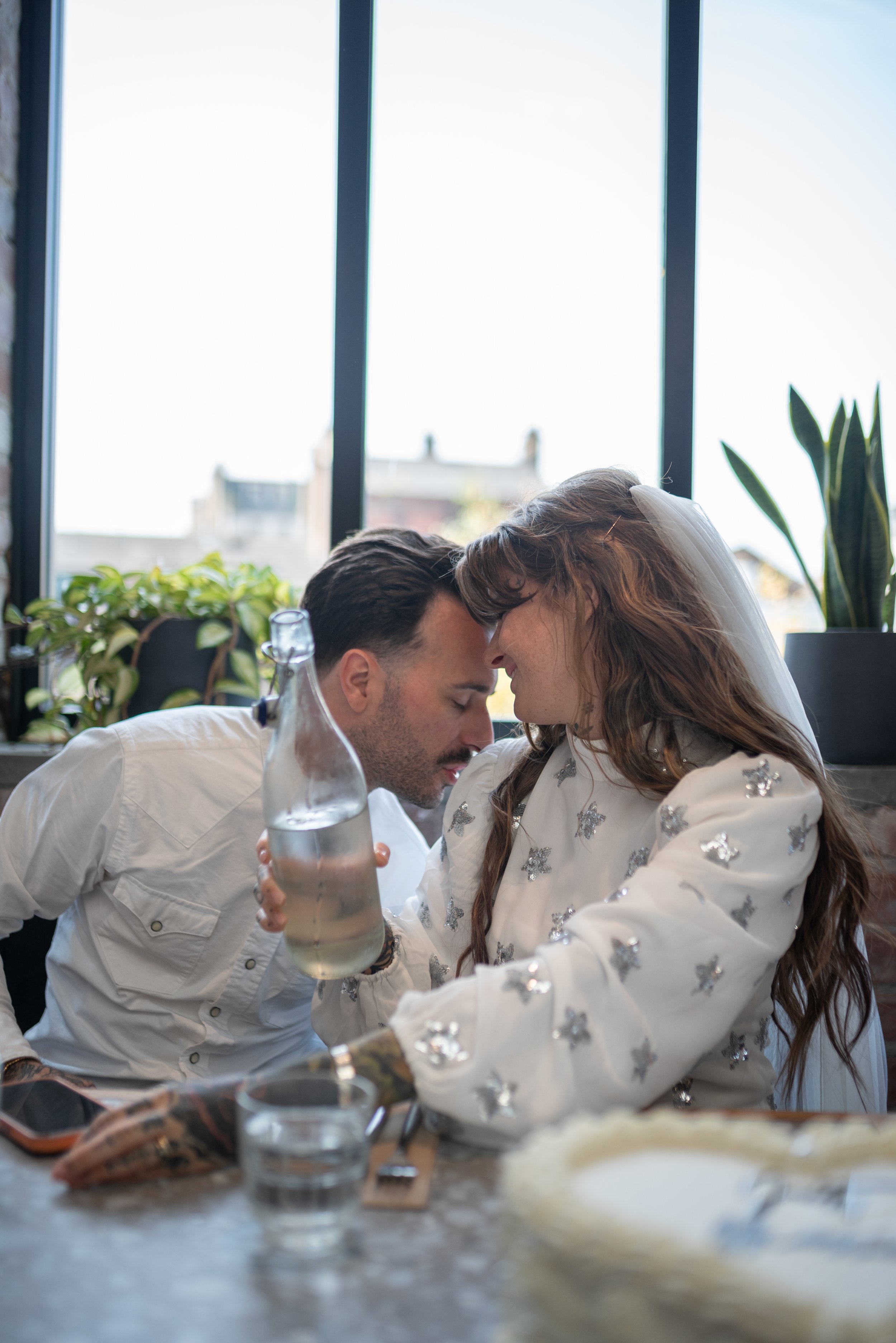 A couple leaning their foreheads together at a table in a bright, modern space with large windows, plants, and a bottle of water.