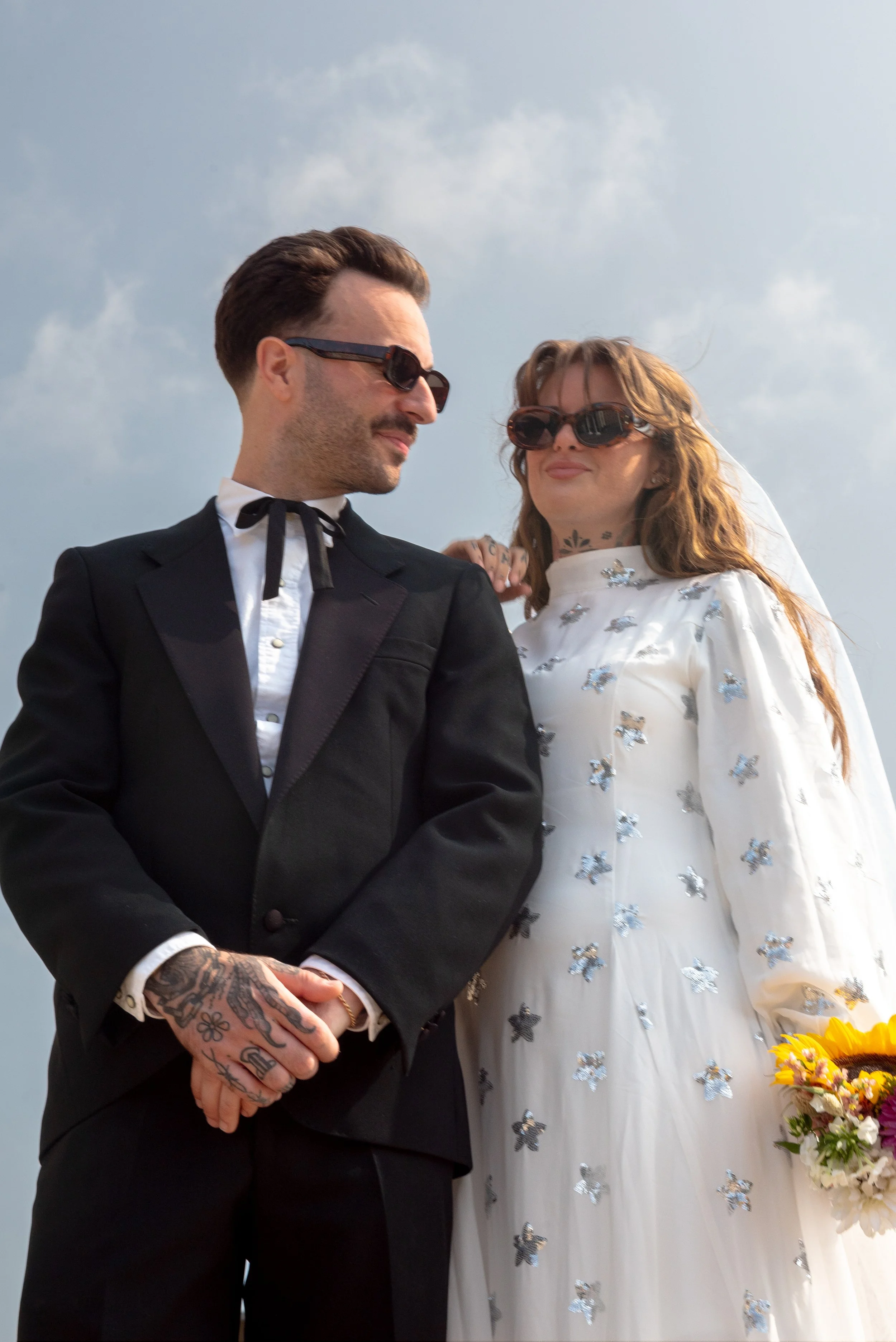 A couple dressed in wedding attire, with the man in a black suit and the woman in a white dress with butterfly embellishments, standing outdoors under cloudy sky, both wearing sunglasses.