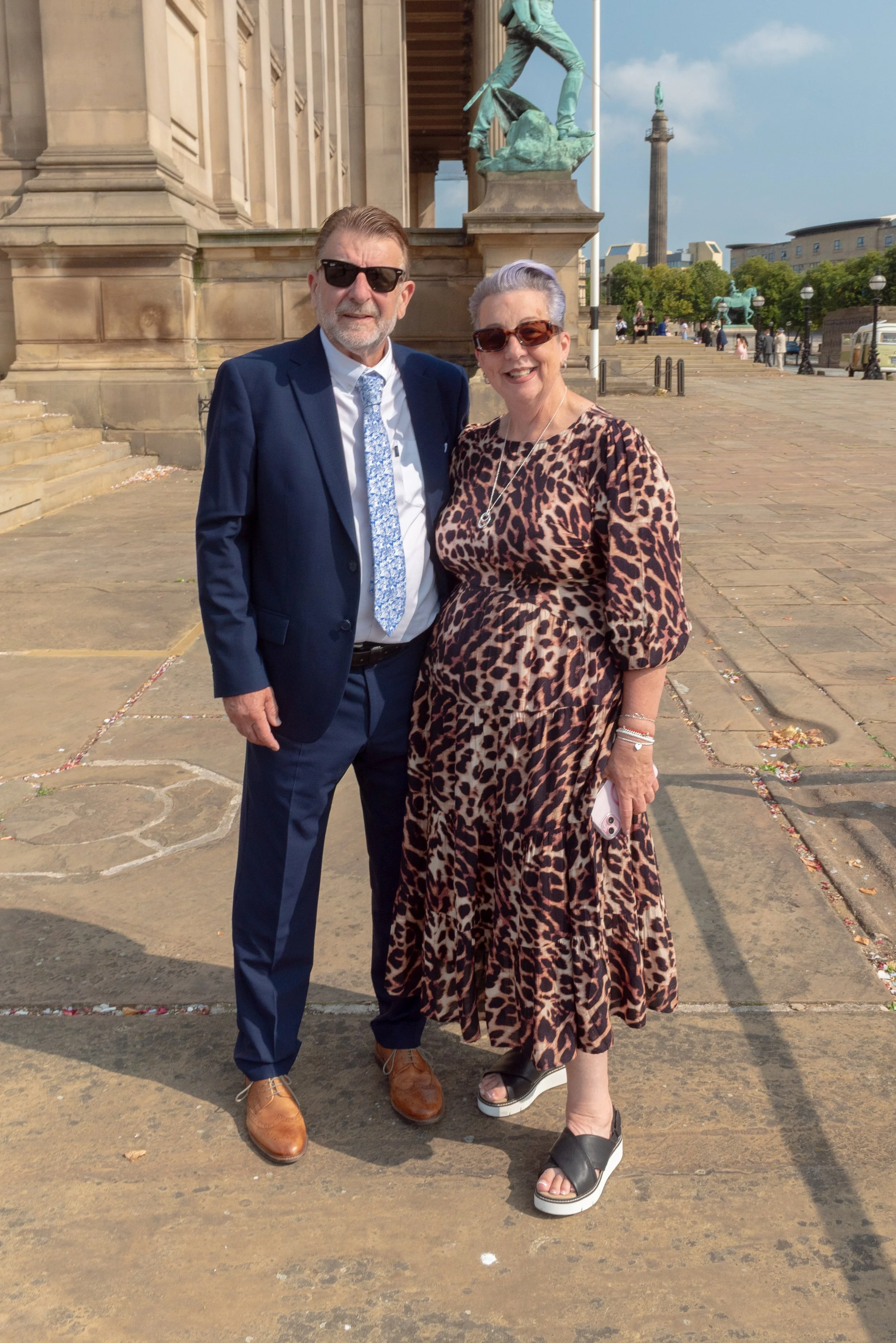 Two people, a man in a blue suit and a woman in a leopard print dress, standing outside in front of a historic building with a large statue and tall column in the background. The weather appears sunny and clear.