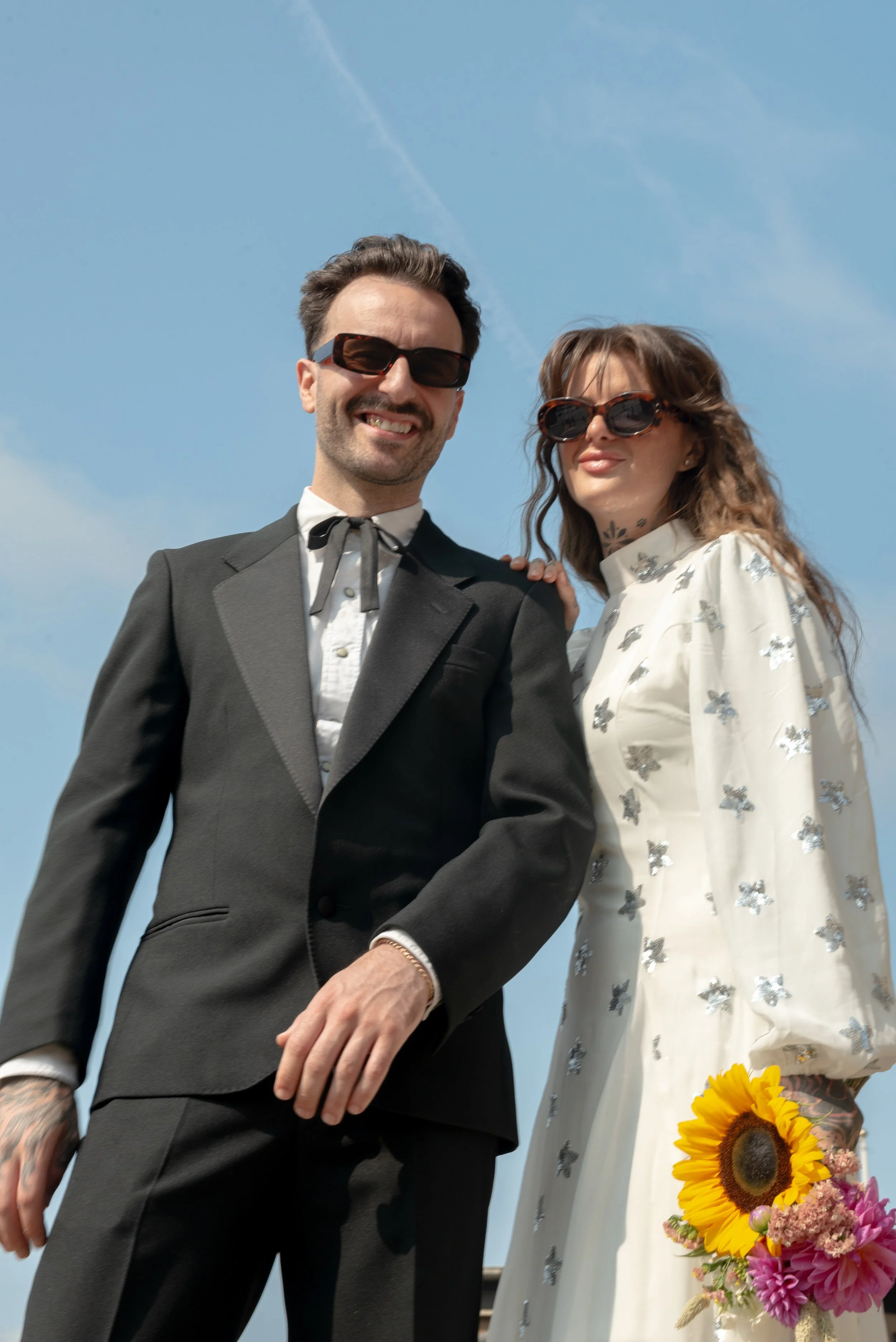 A smiling man and woman dressed in vintage style clothing standing outdoors against a bright blue sky, both wearing sunglasses. The woman holds a bouquet of flowers including a sunflower and pink blooms.