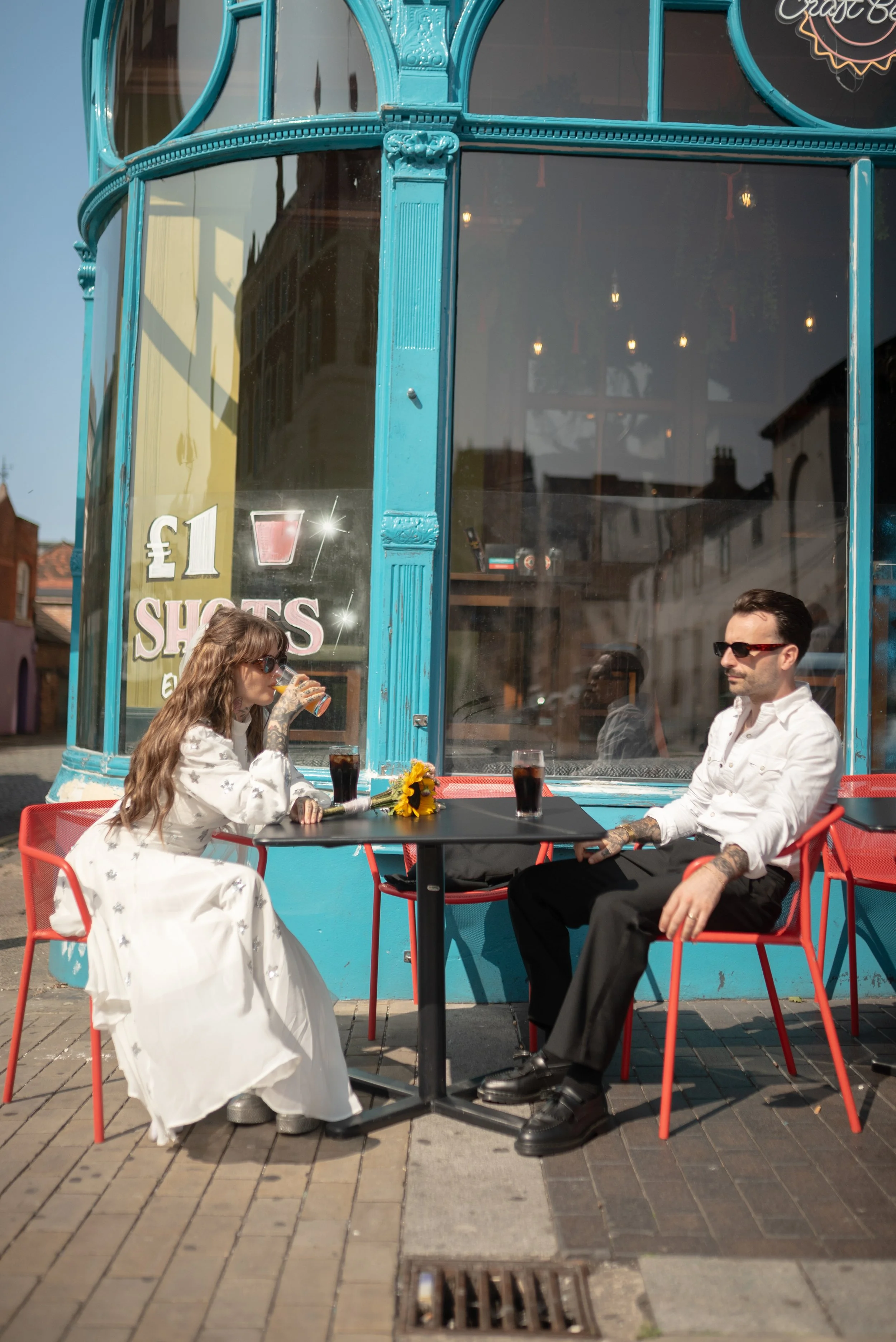 A man and woman are seated outside a restaurant at a small table on a sunny day. The woman is drinking from a glass, and the man is sitting with his hands on the table. The woman has long wavy hair and sunglasses, and the man has tattoos on his arms,